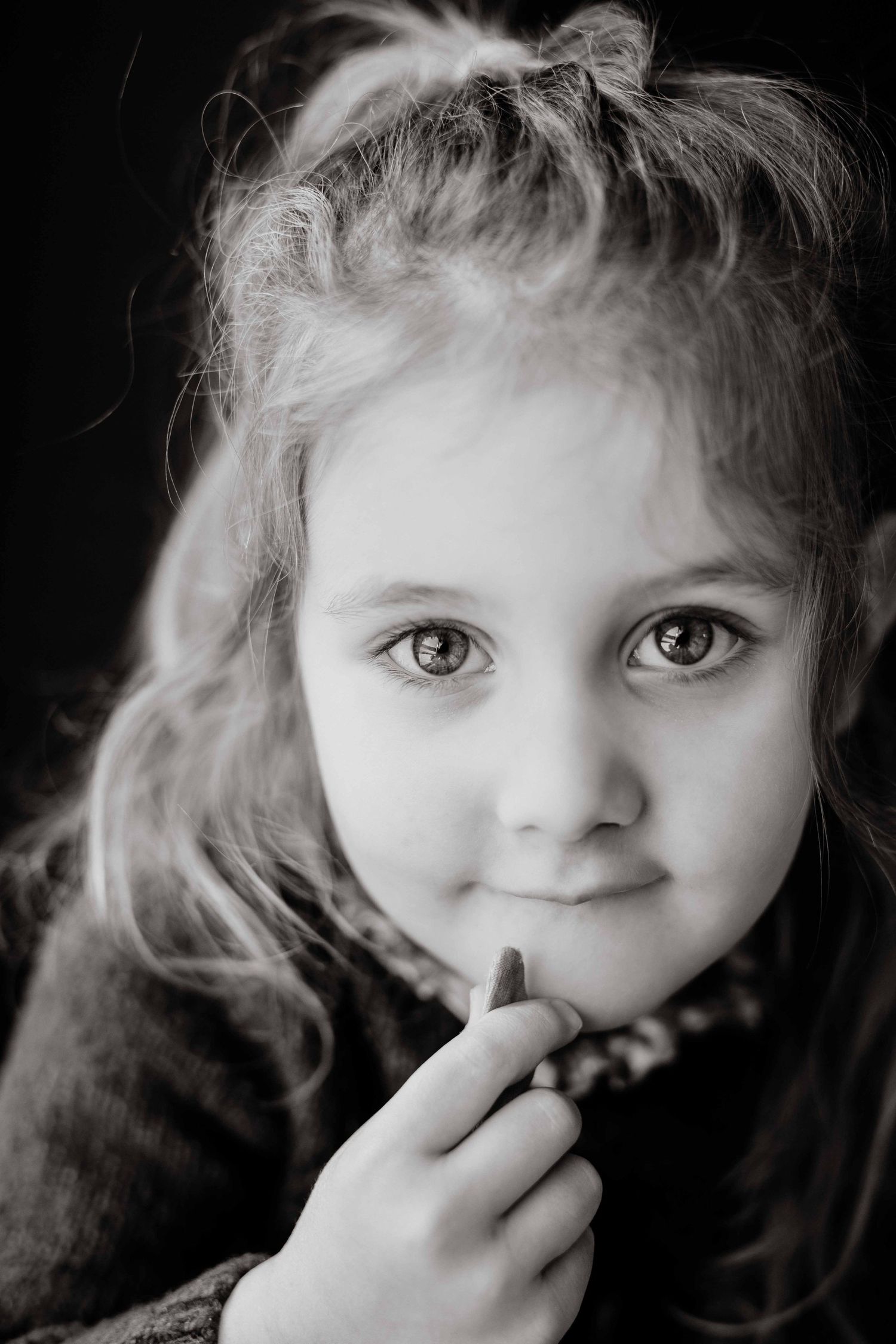 Black and white portrait of a young girl with messy updo hair holding a crayon under her chin with a sweet smile.