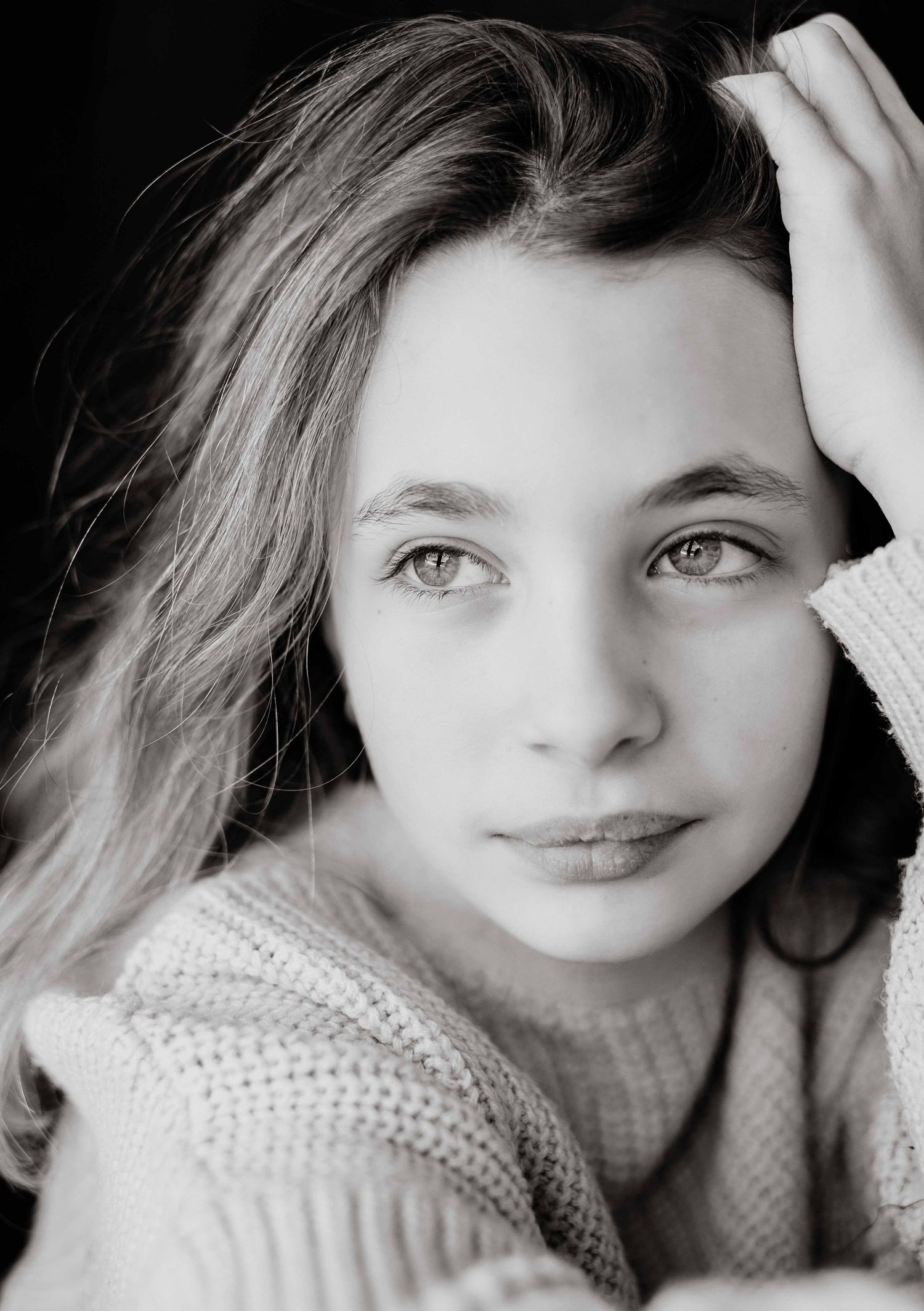 Black and white portrait of a young girl with long wavy hair resting her head on her hand with a calm, pensive gaze.