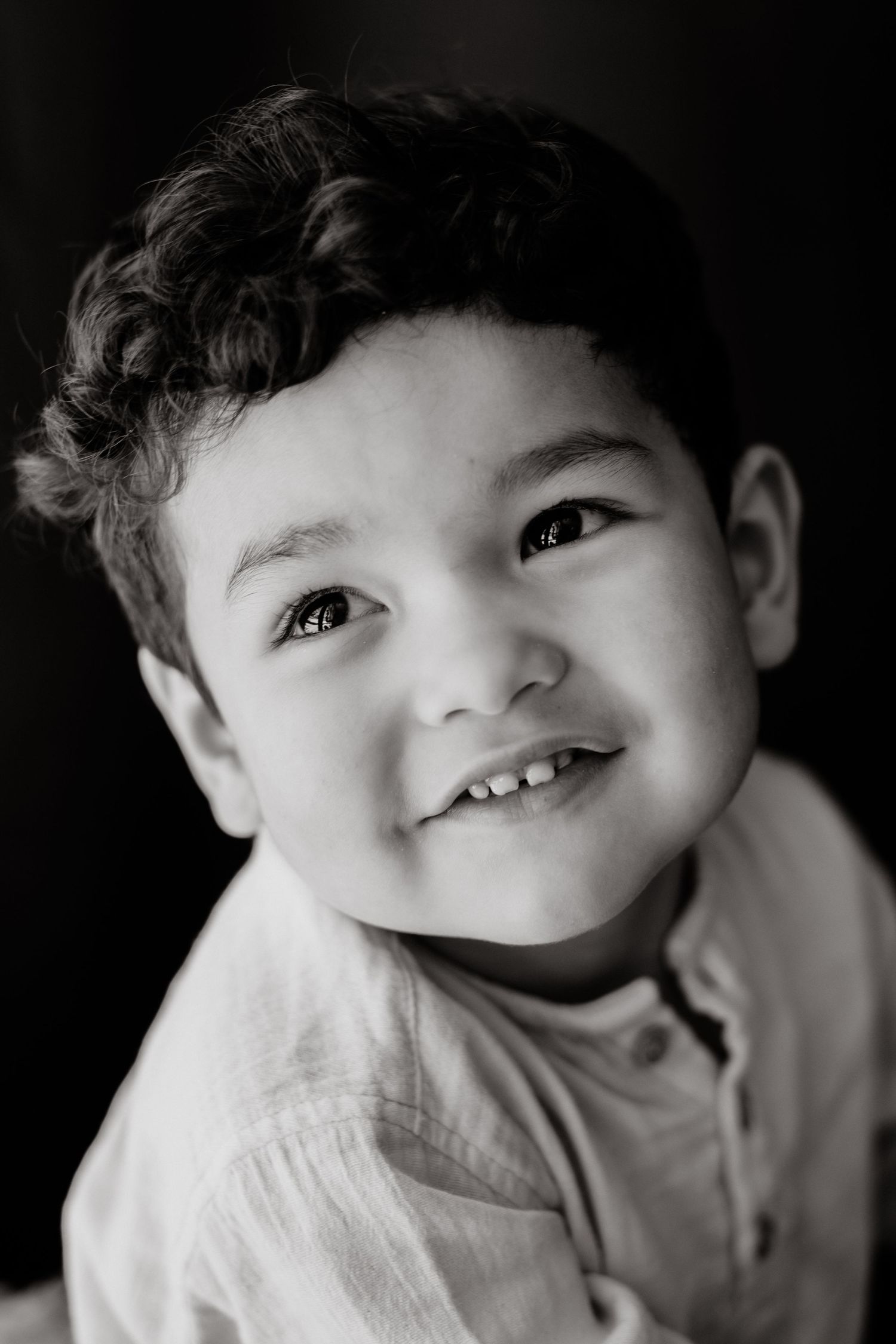 Black and white portrait of a curly-haired little boy in a linen shirt smiling and looking up joyfully.