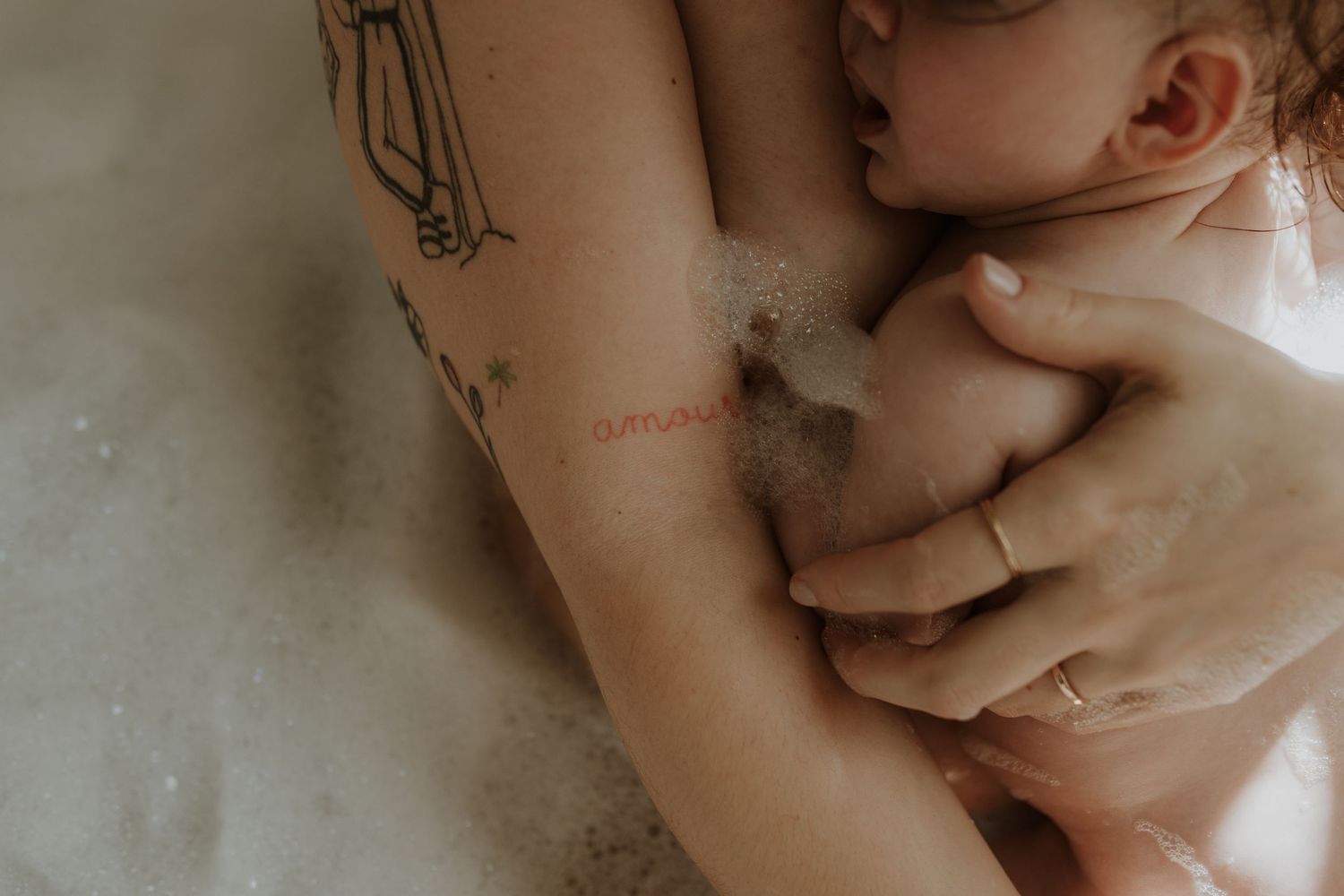 Close-up of a tattooed parent holding a baby in a bubble bath, with a hand gently cradling the infant.