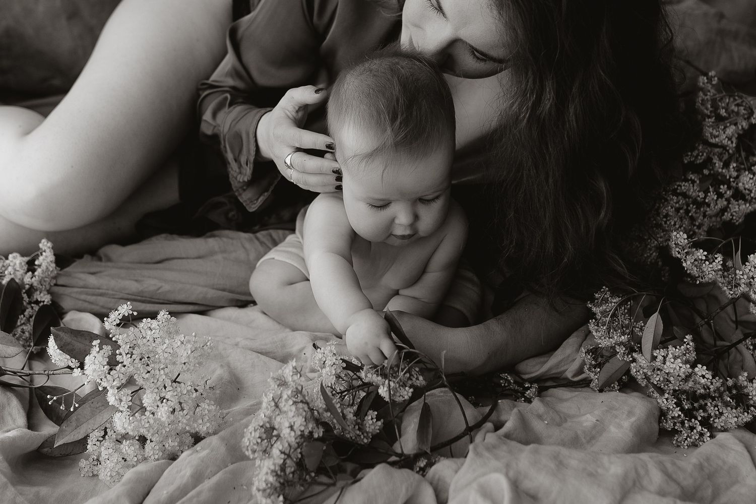 Black and white photo of a mother lying beside her baby, who sits on a blanket surrounded by wildflowers in a tender moment.