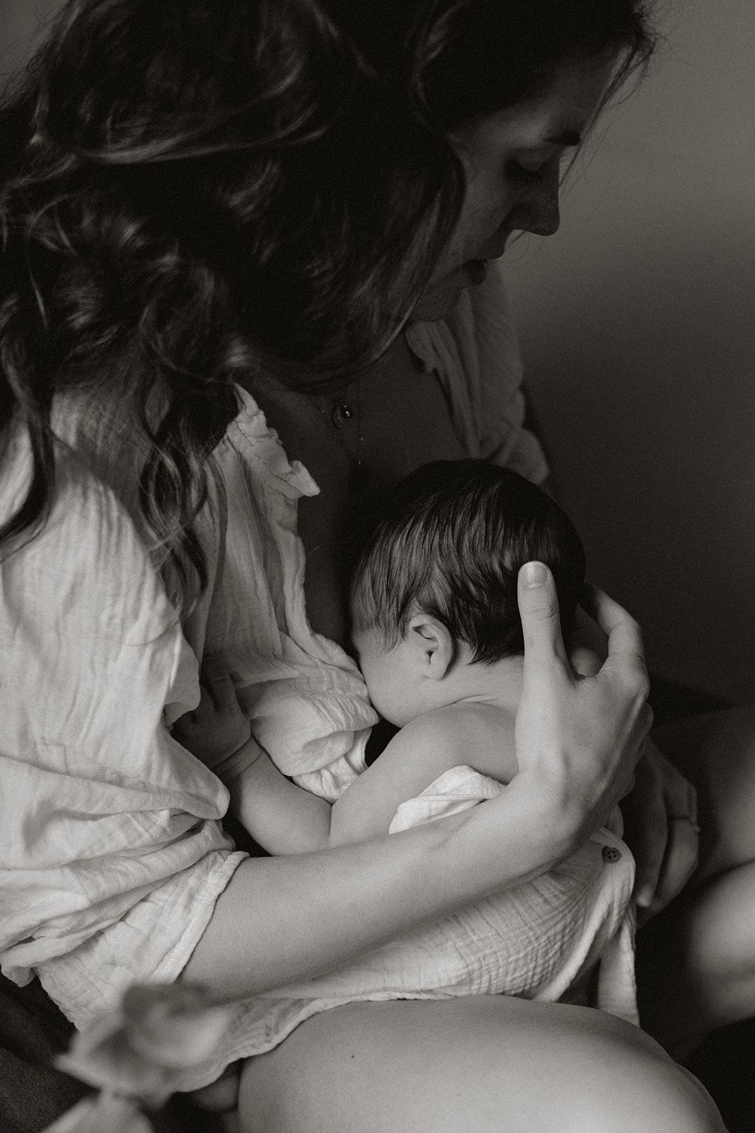 Black and white photo of a mother with wavy hair gently cradling and breastfeeding her newborn baby.