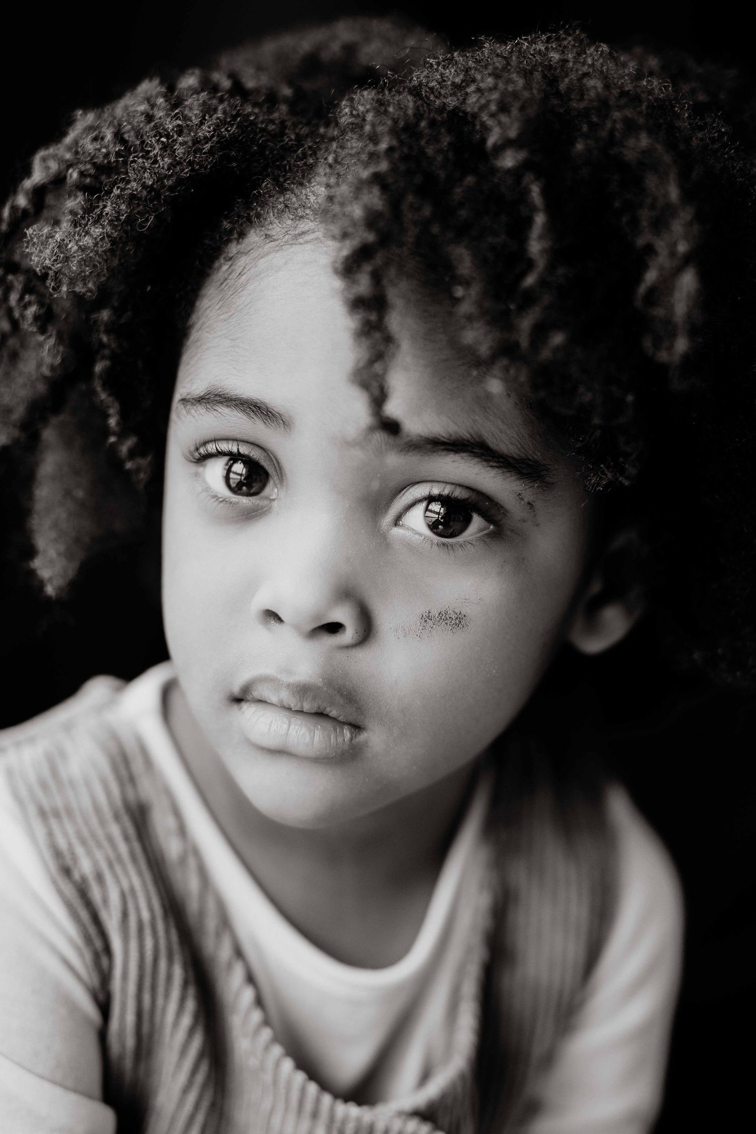 Black and white portrait of a young girl with curly hair and expressive eyes looking solemnly at the camera.