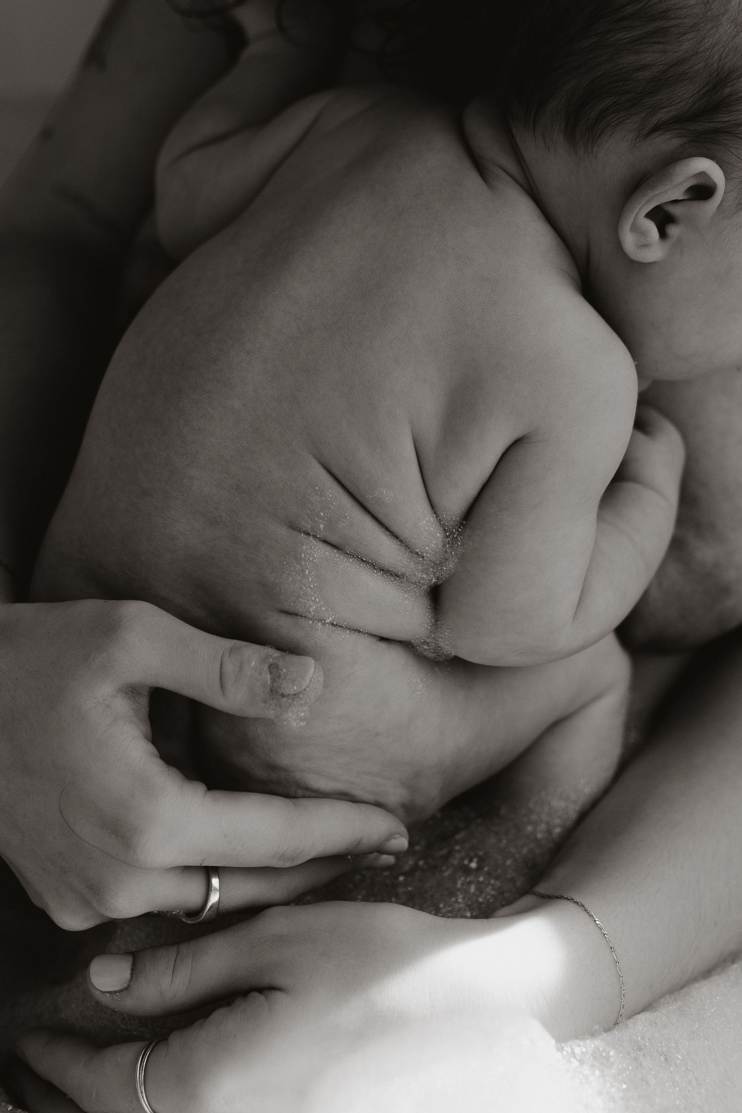 Black and white close-up of a newborn baby held gently in a parent's hands, showing a wedding ring and tiny ear.