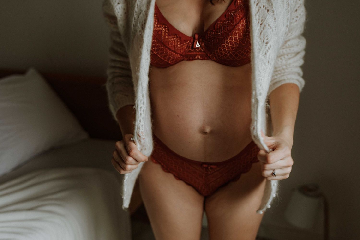 Pregnant woman in red lace lingerie and a cream knit cardigan standing beside a bed in soft natural light, her baby bump visible.