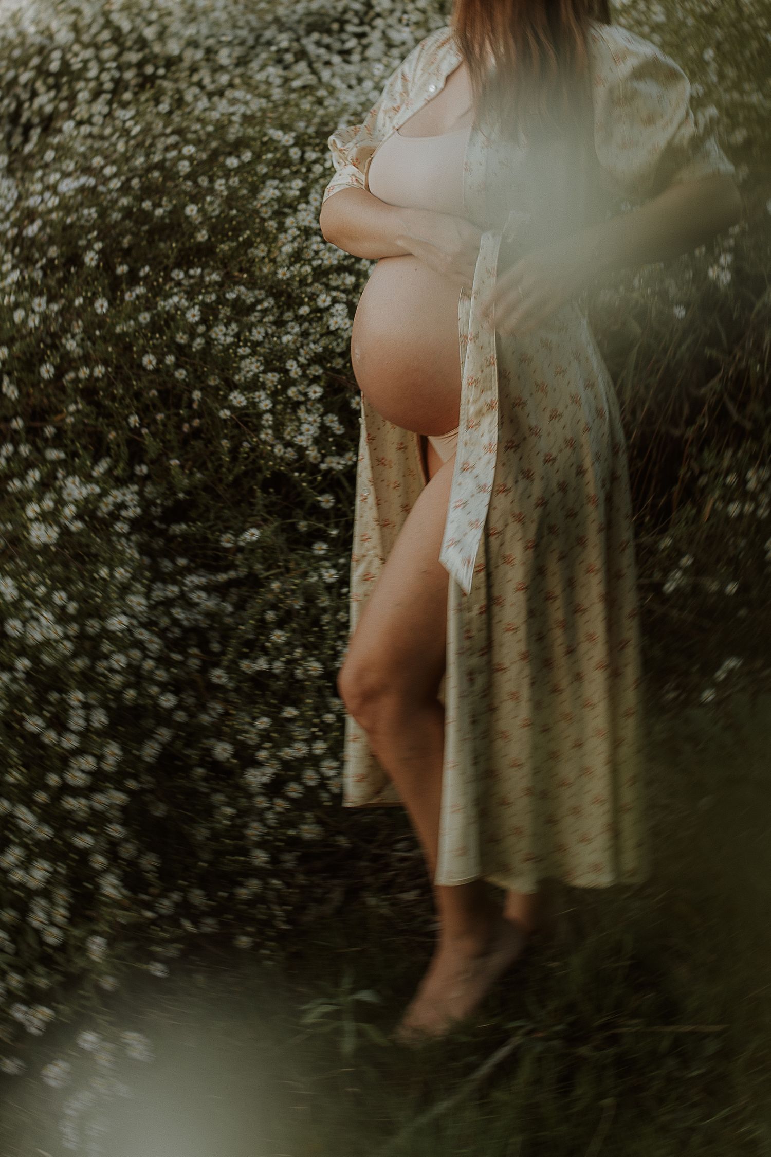 Pregnant woman in a floral robe cradles her bare belly while standing barefoot beside a field of wild white daisies.