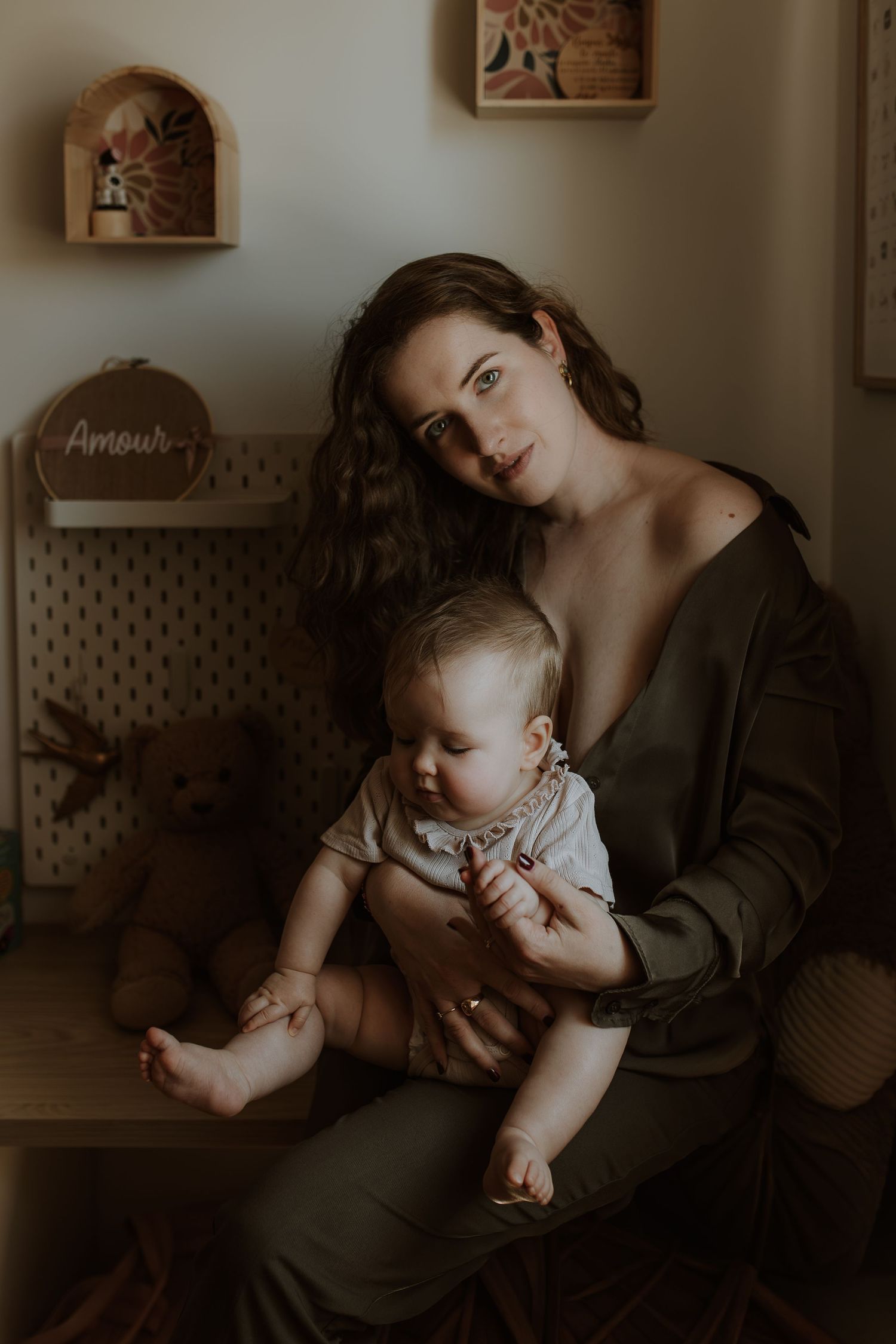 A mother with wavy dark hair sits holding her smiling baby on her lap in a cozy nursery with a teddy bear and wall shelves.