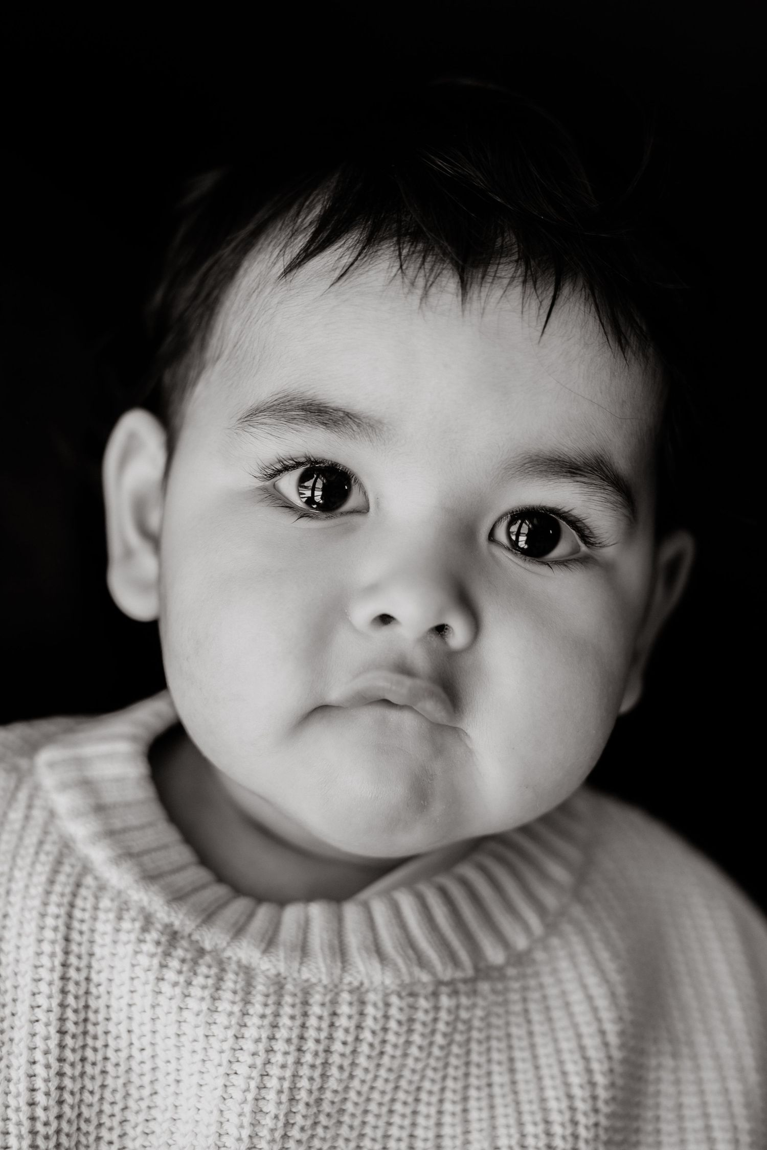 Black and white portrait of a chubby-cheeked toddler in a knit sweater with a serious pouty expression.