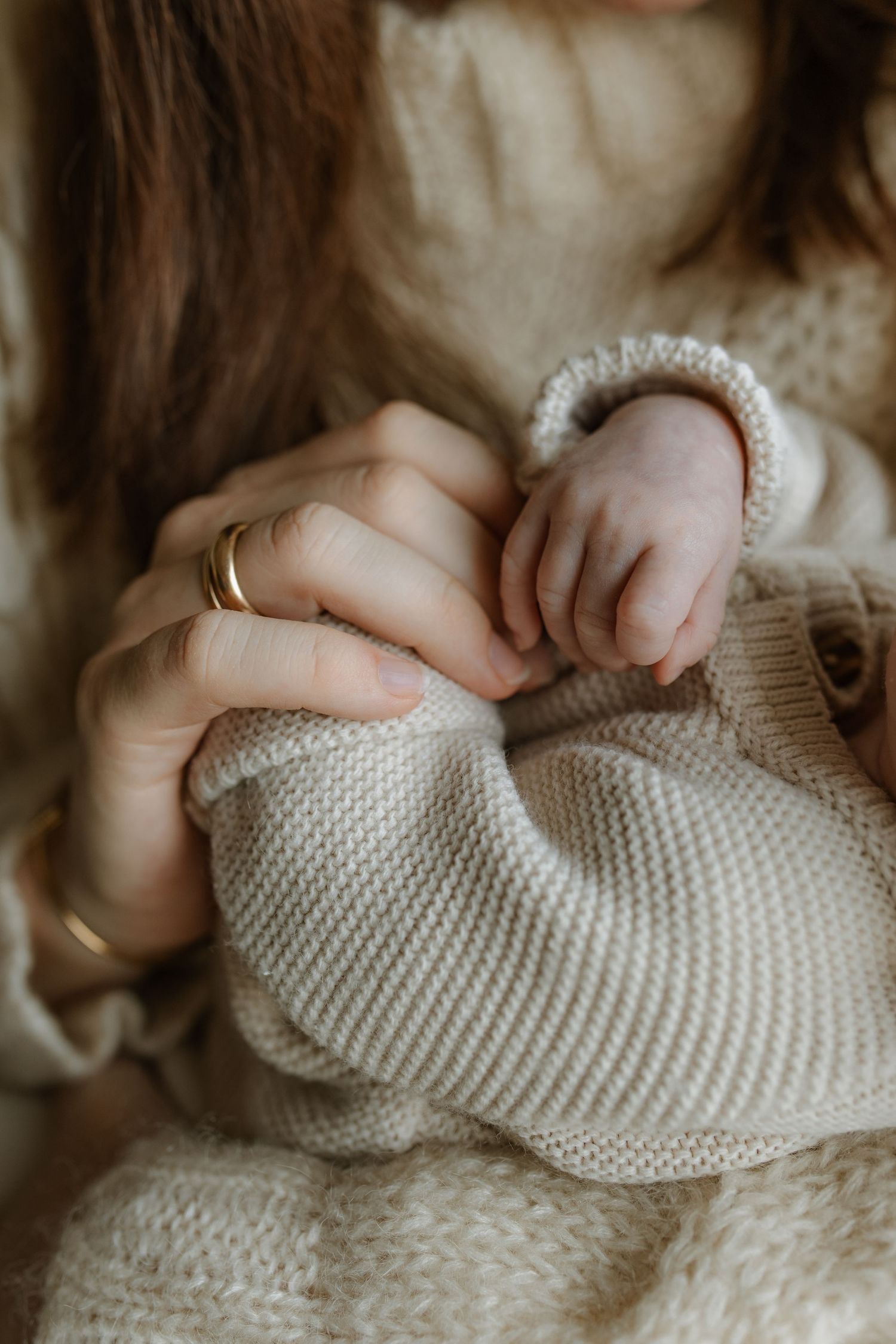 A mother's hands wearing gold rings gently hold a newborn baby's tiny hand wrapped in a cozy cream knit sweater.