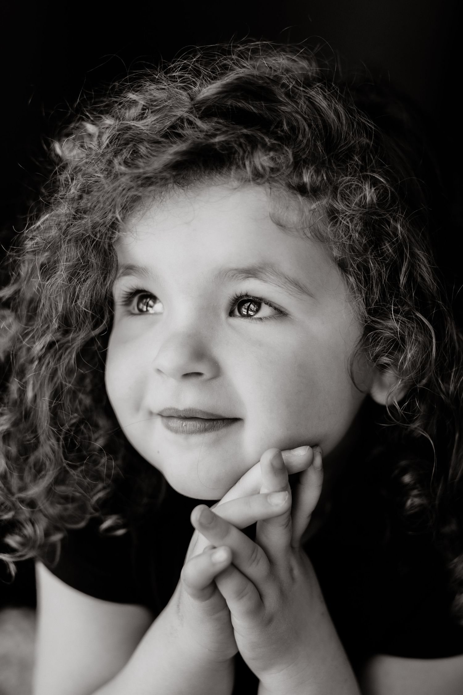Black and white portrait of a smiling girl with curly hair resting her chin on clasped hands and gazing upward.