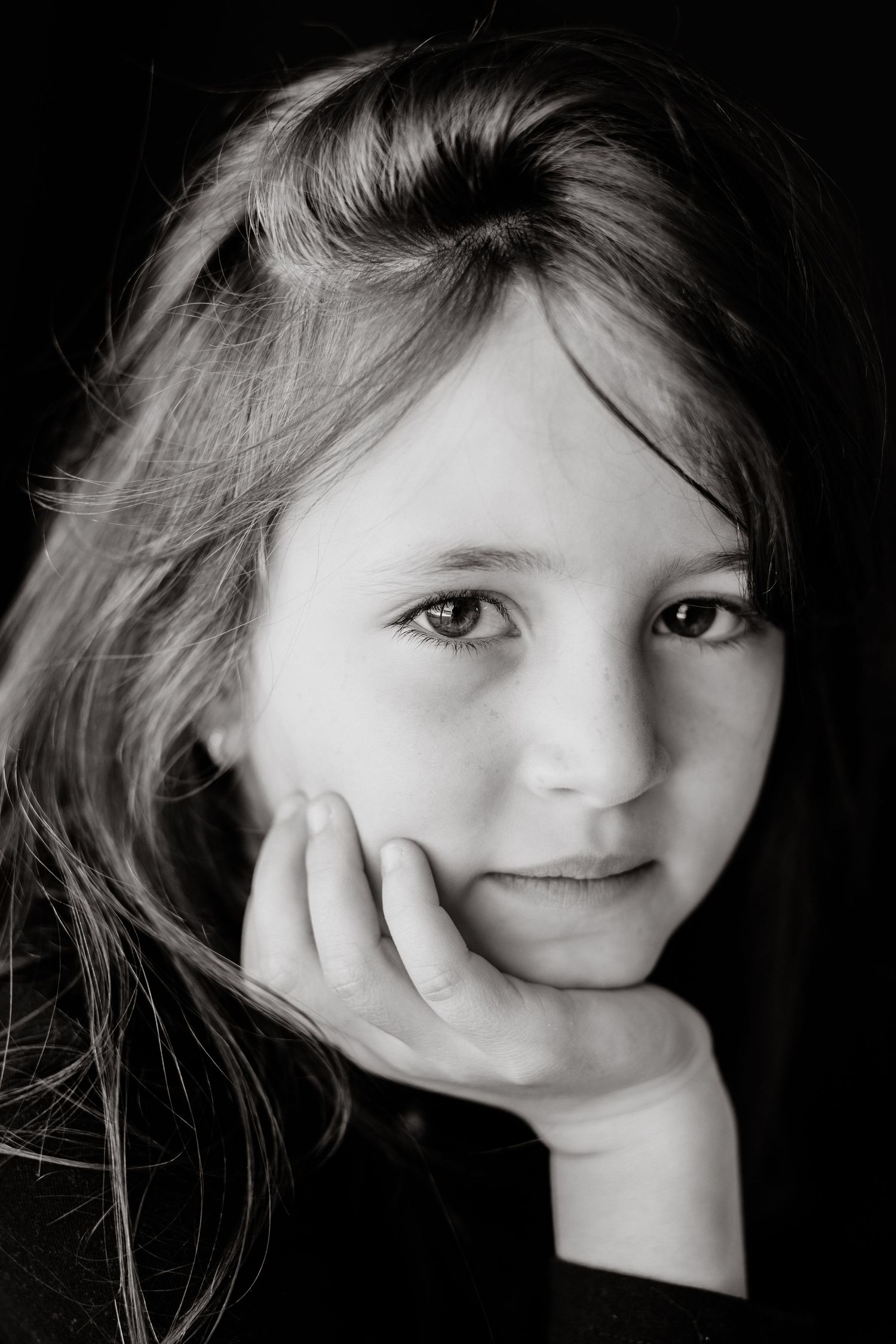 Black and white portrait of a girl with long hair resting her chin on her hand and looking softly at the camera.