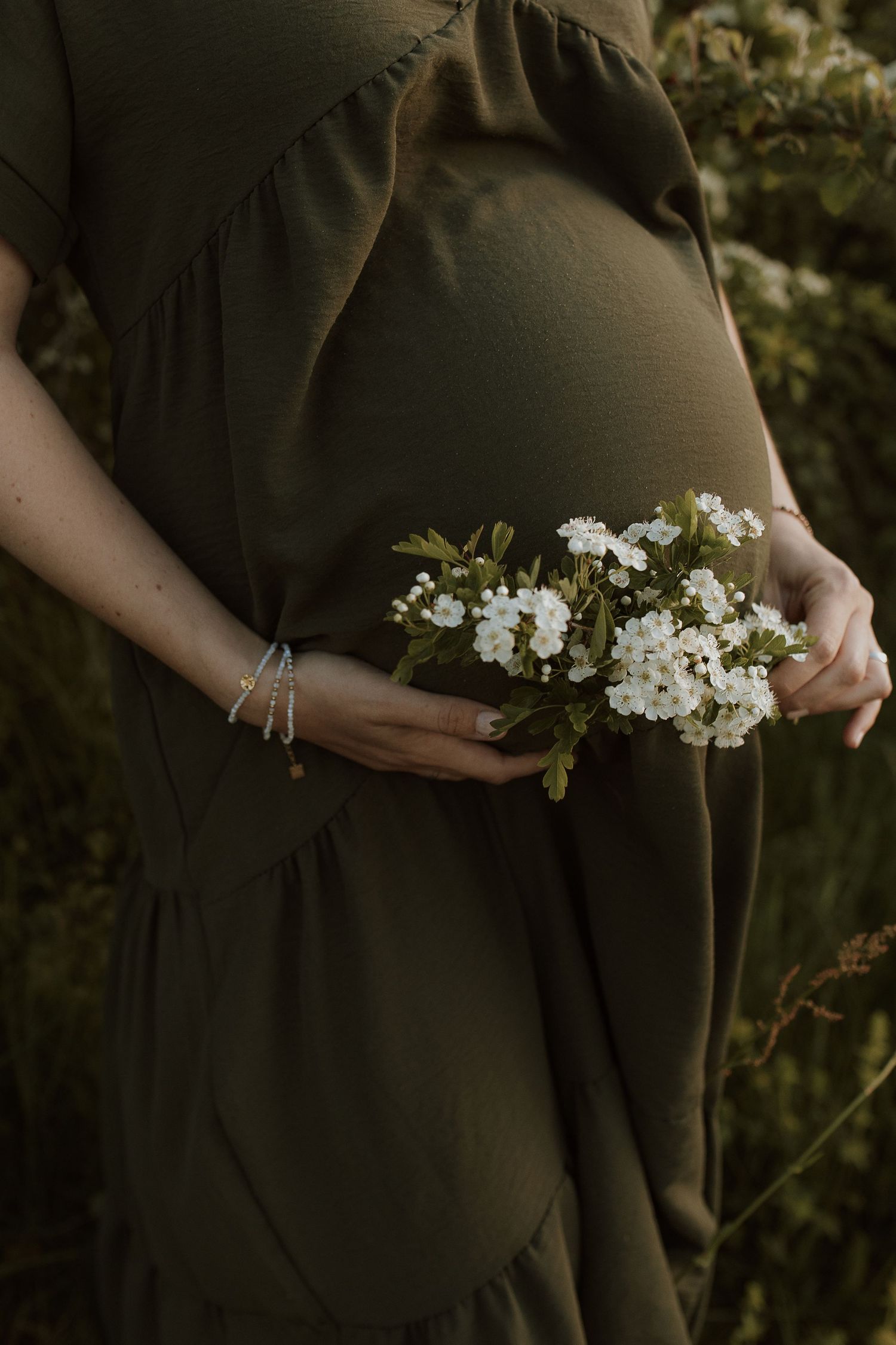Pregnant woman in an olive green dress holding a bouquet of small white flowers against her belly outdoors.