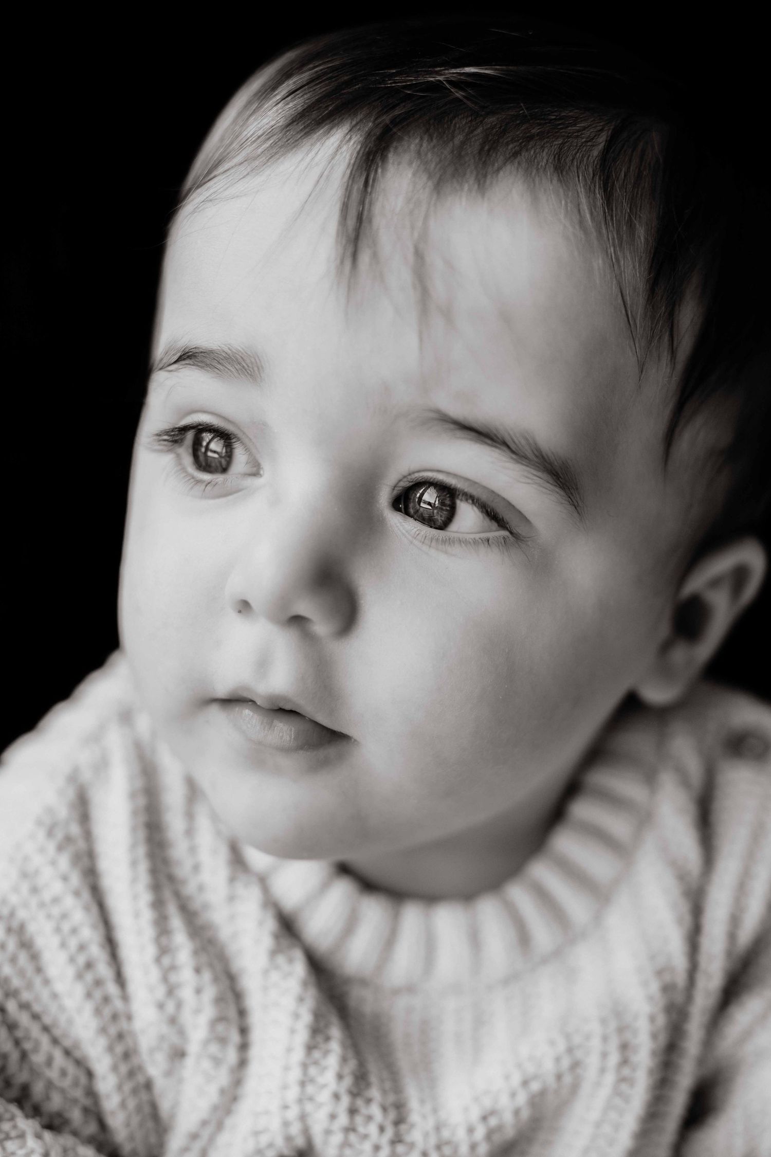 Black and white close-up portrait of a baby boy in a chunky knit sweater gazing thoughtfully to the side.