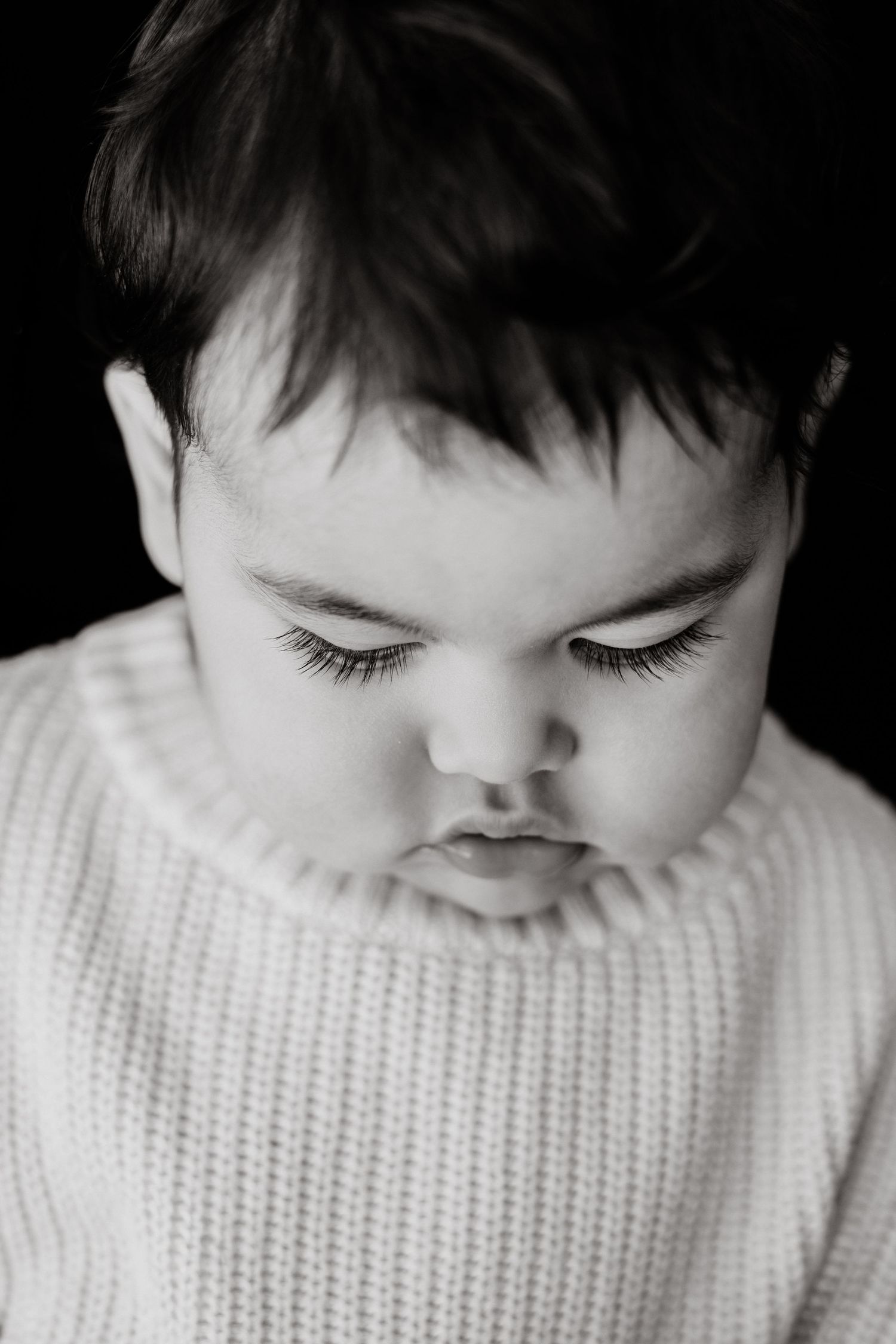 Black and white portrait of a baby with dark hair looking down, wearing a chunky knit sweater against a dark background.