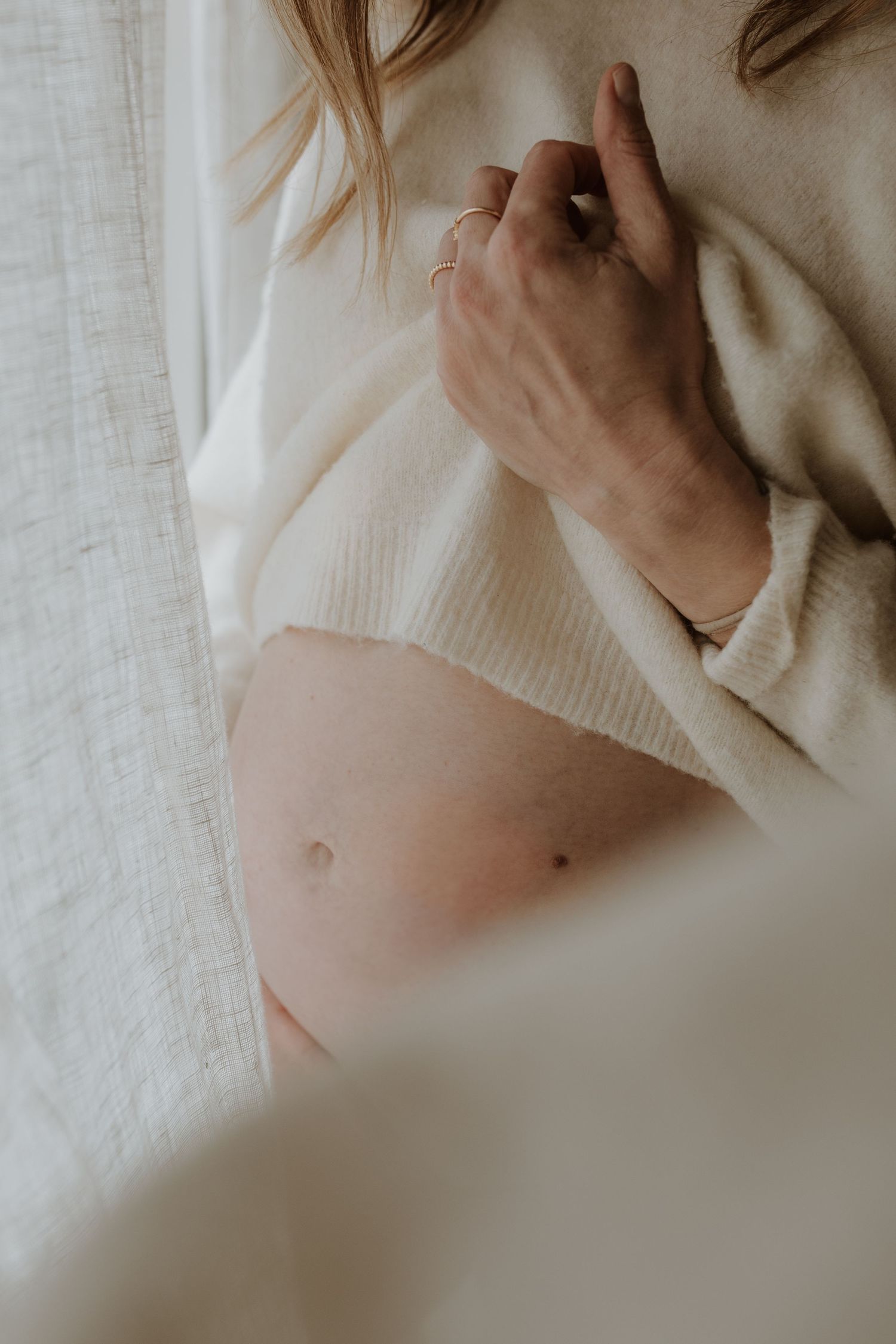 Close-up of a pregnant belly with a woman's ringed hand resting on her cream knit sweater beside a linen curtain.