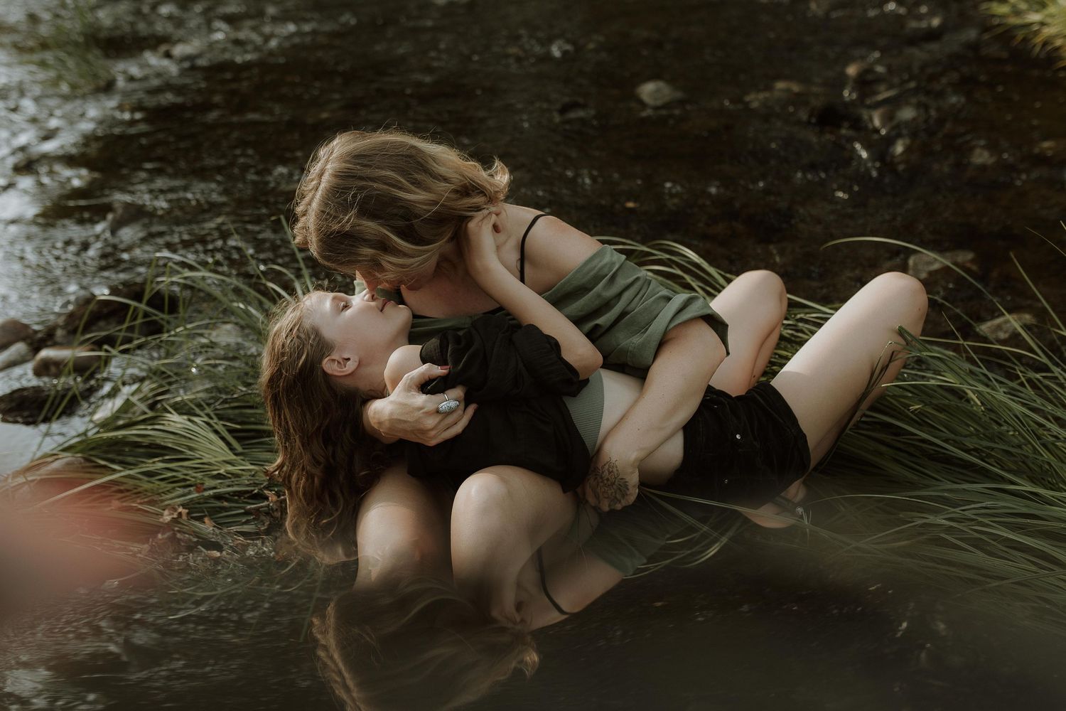 Two women share a tender moment lying in shallow water among tall grass in a moody, natural outdoor portrait.