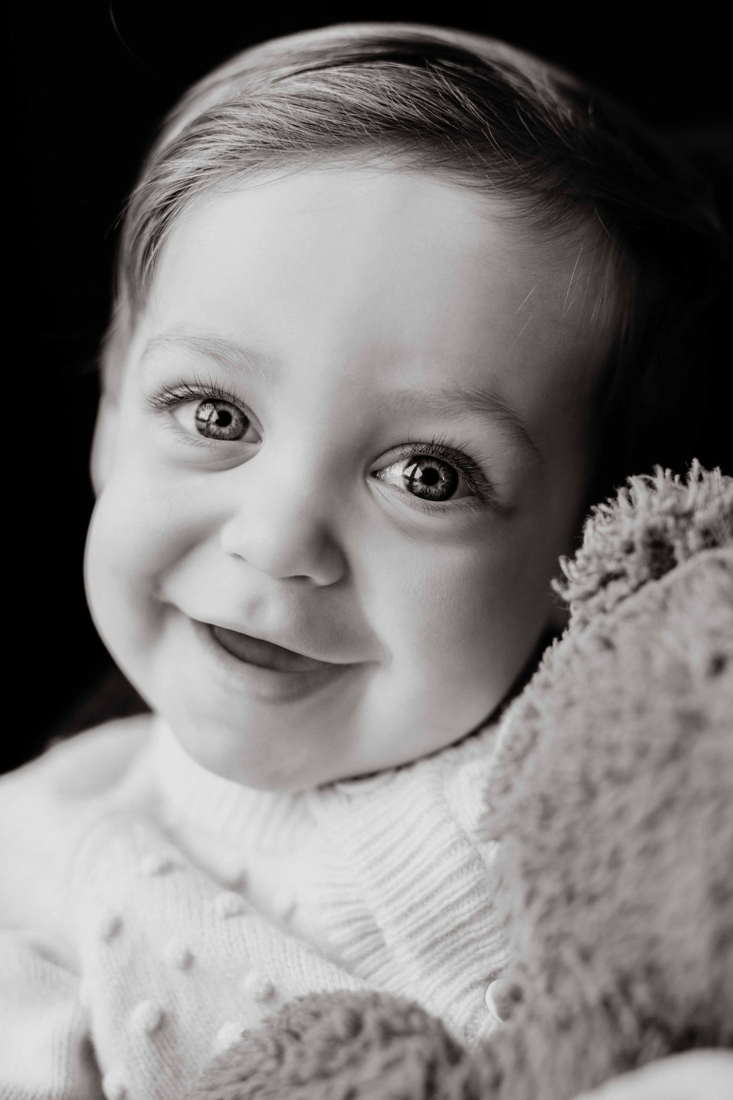 Black and white portrait of a smiling baby with big expressive eyes snuggling a plush teddy bear.