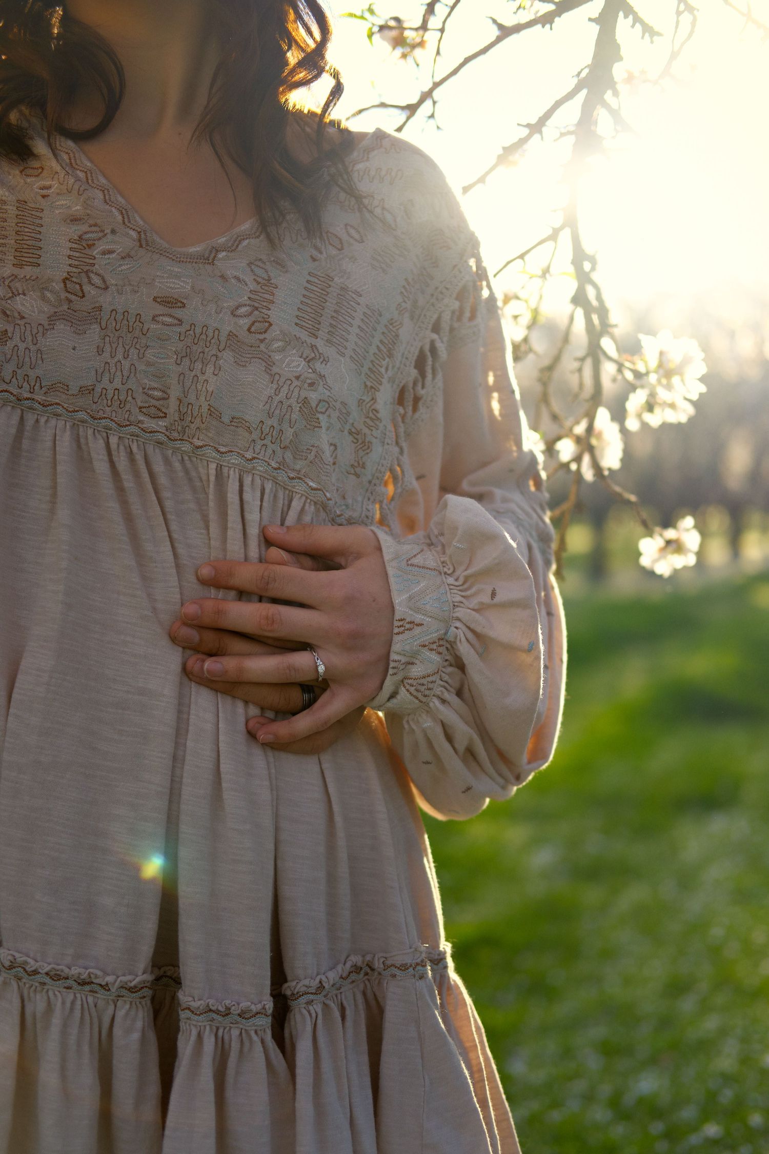 A woman in a cream boho dress rests her hand on her stomach, wearing a ring, with spring blossoms and golden sunlight behind her.