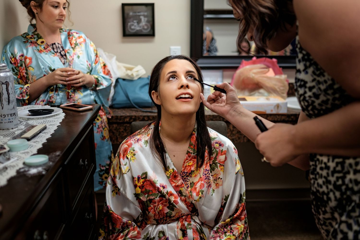Bride in a floral satin robe has makeup applied by an artist while a bridesmaid in a matching robe looks on during wedding prep.