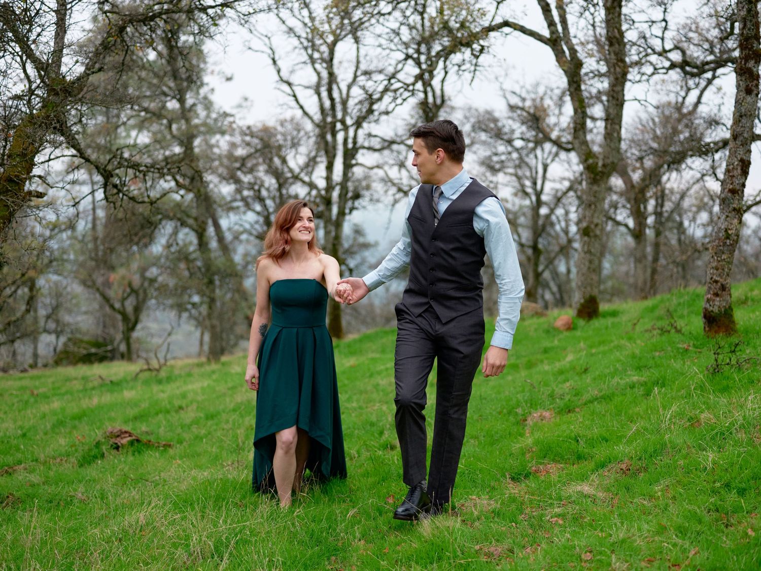 A couple holds hands and walks across a green grassy hillside, the woman in an emerald dress and the man in a dark vest.