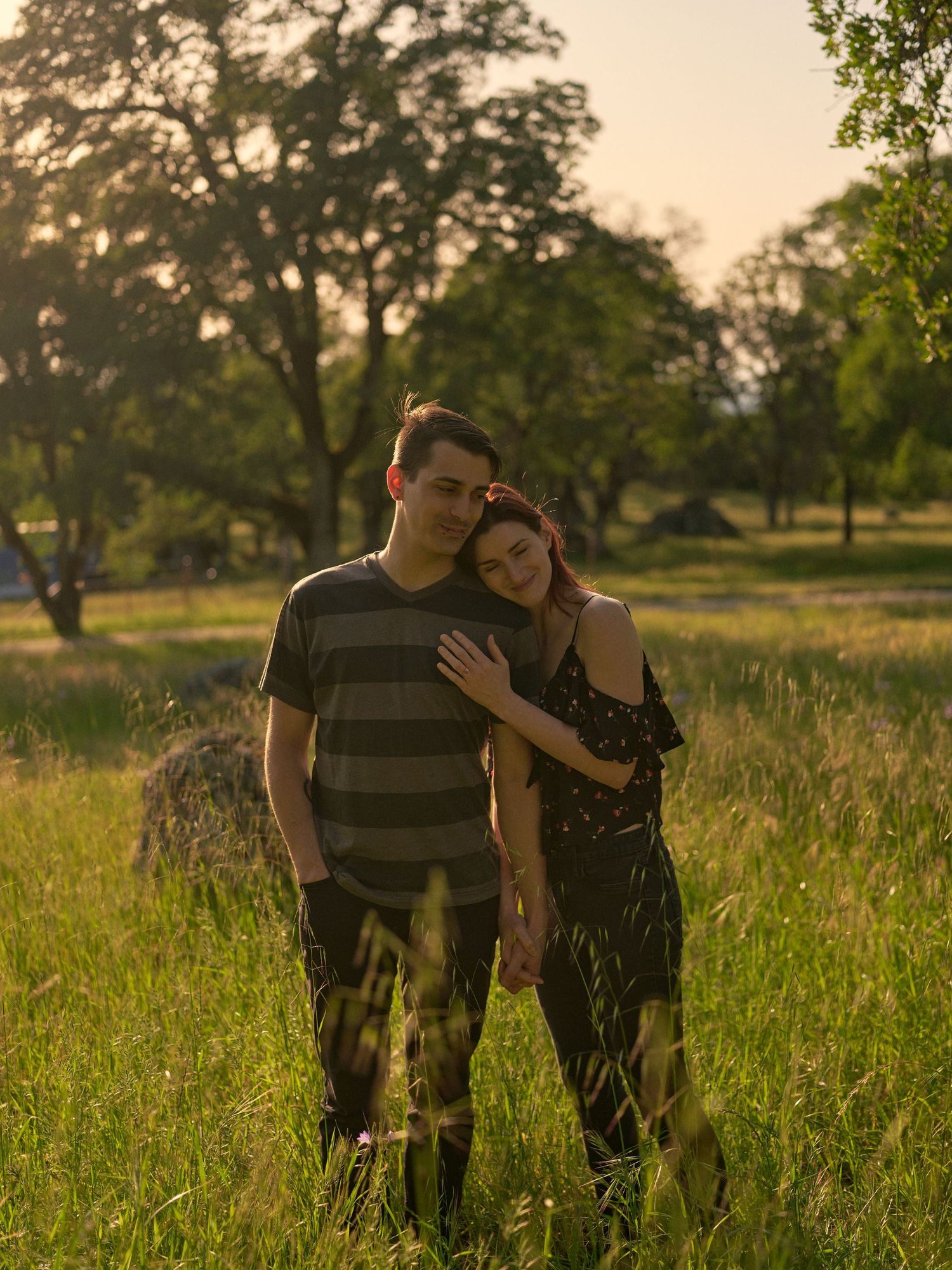 A couple stands embracing in a tall grassy field at golden hour, the woman leaning her head on the man's shoulder among oak trees.