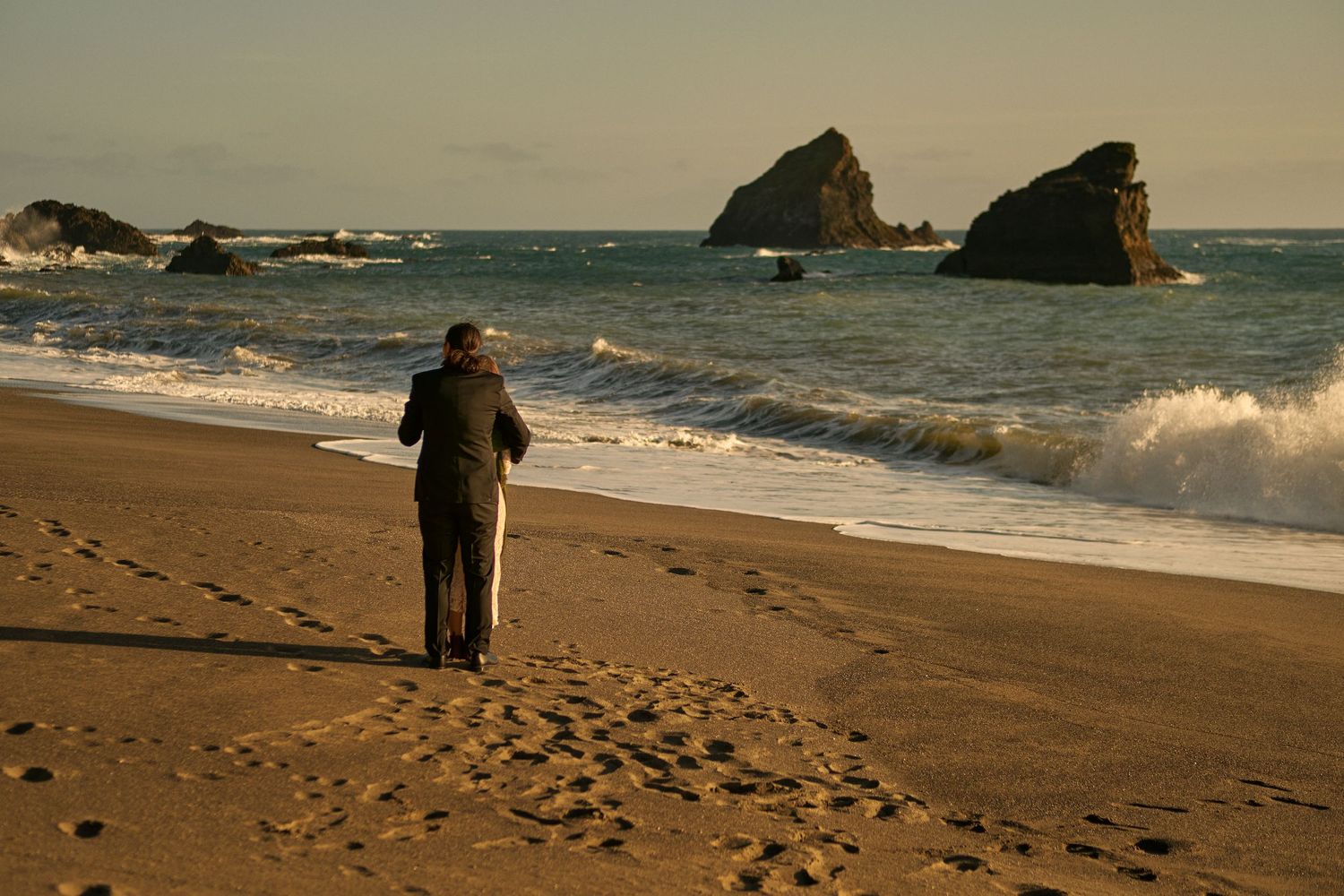Couple in wedding attire stands on a sandy beach at sunset with waves and rocky sea stacks in the background.