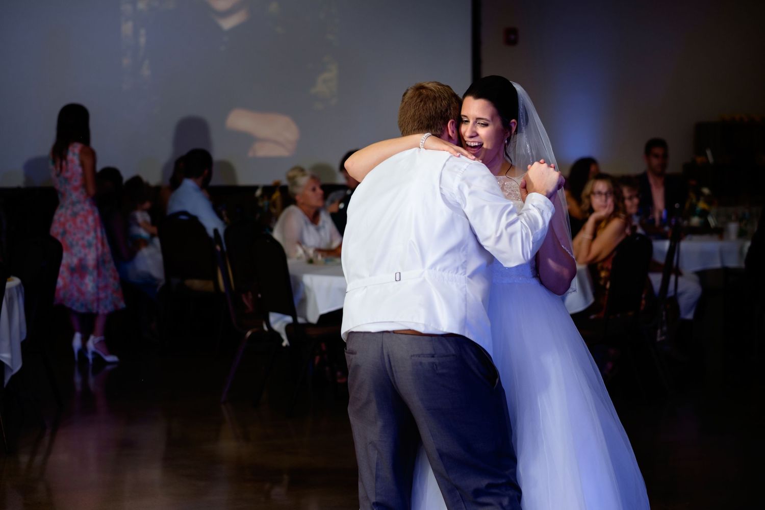 A bride in a white gown and veil embraces a groom in a white shirt during a wedding reception dance as guests look on.