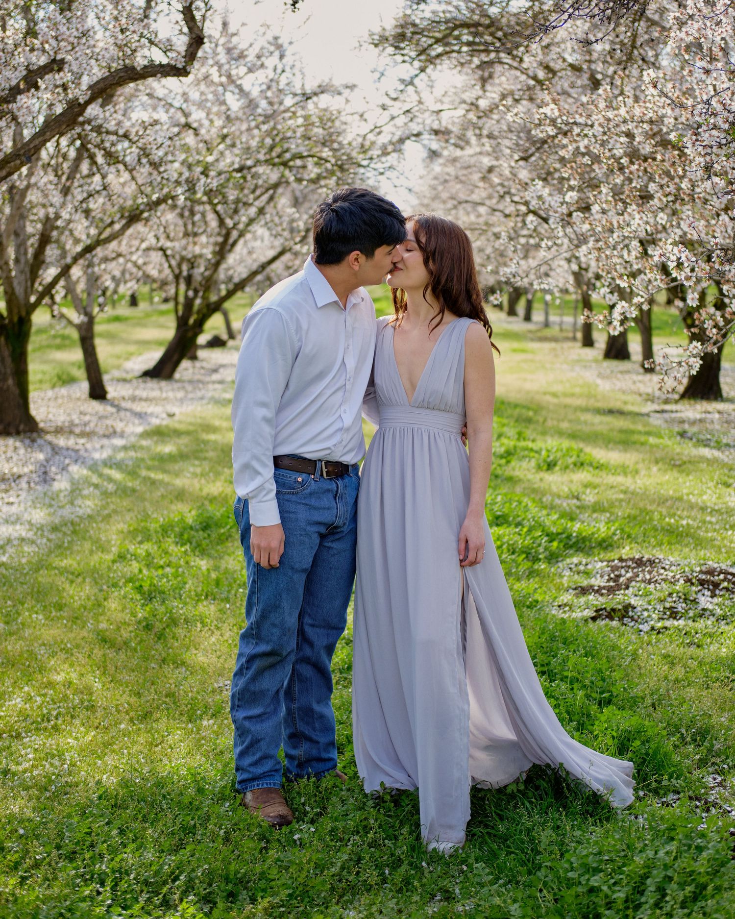 A couple in an orchard of blooming almond trees share a close moment, the woman in a long gray gown and the man in jeans.