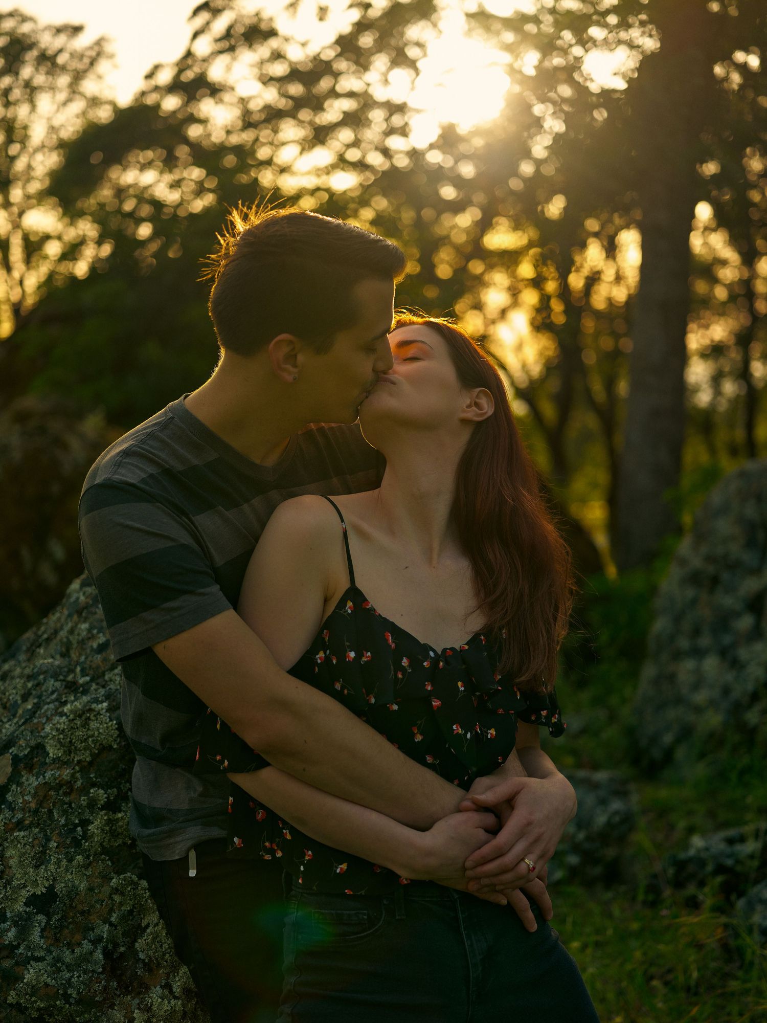 A couple kiss at sunset in a forest as golden sunlight filters through the trees, with the man holding the woman close.