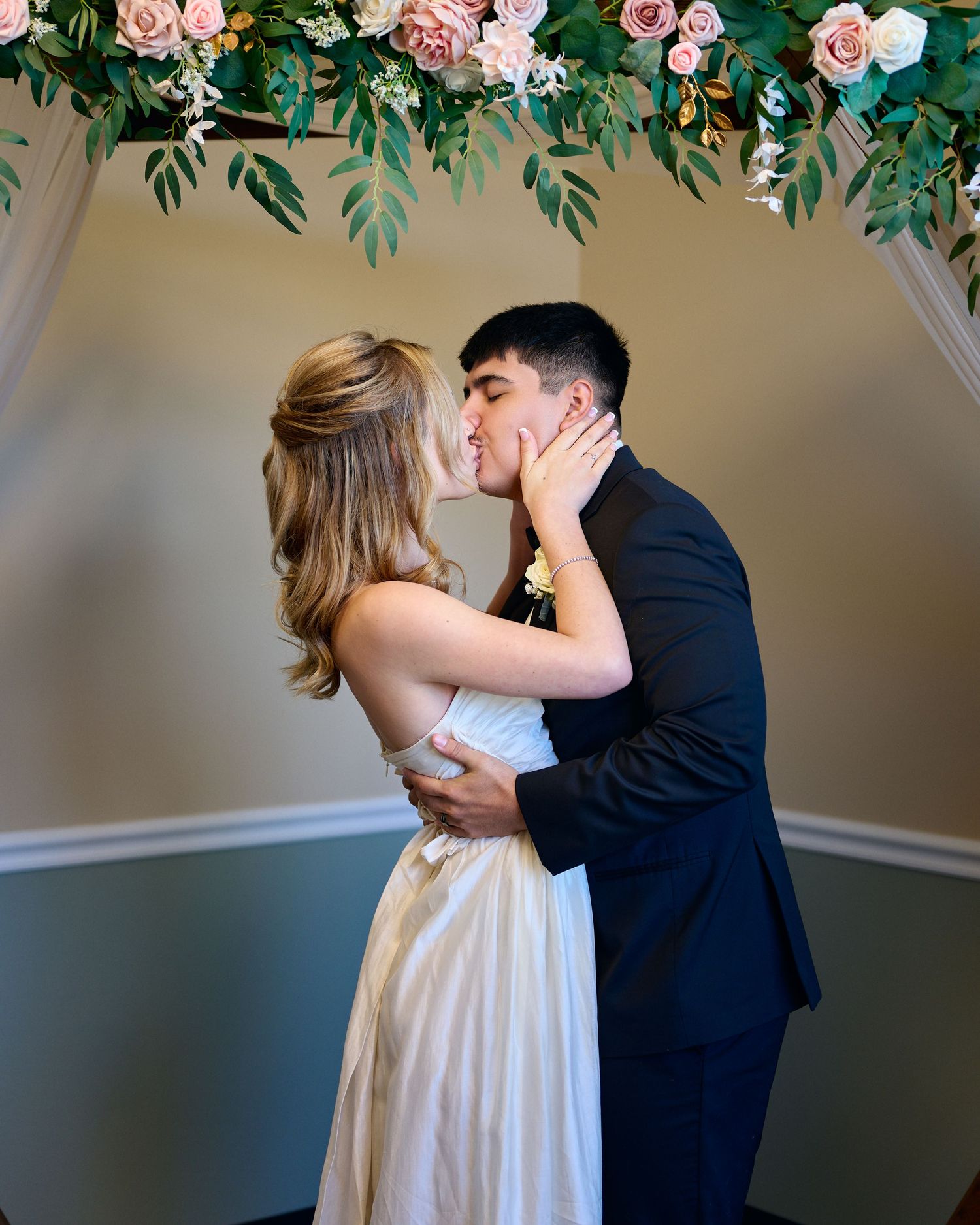 A bride and groom share a kiss beneath a floral arch of pink roses and greenery during their wedding ceremony.