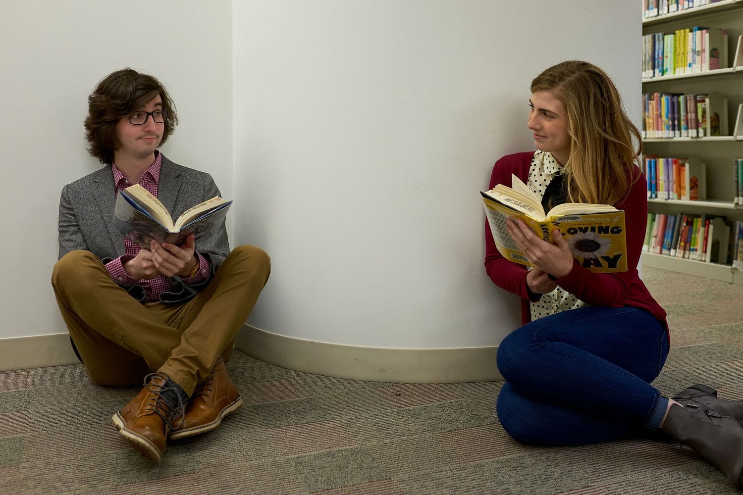 A young man in glasses and a young woman sit on a library floor reading books and smiling at each other near tall bookshelves.
