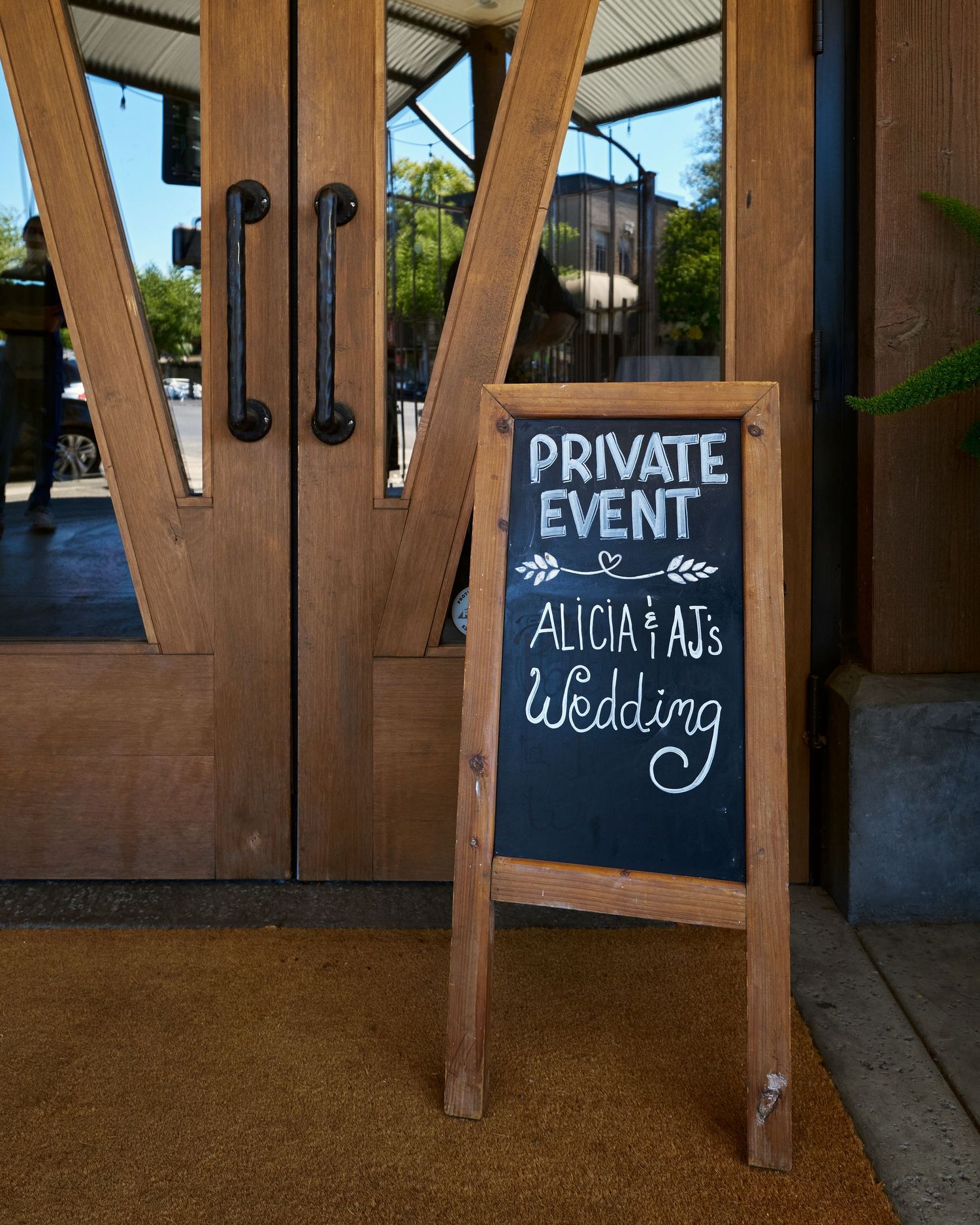 A chalkboard sign reading Private Event, Alicia and AJ's Wedding stands beside rustic wooden double doors.