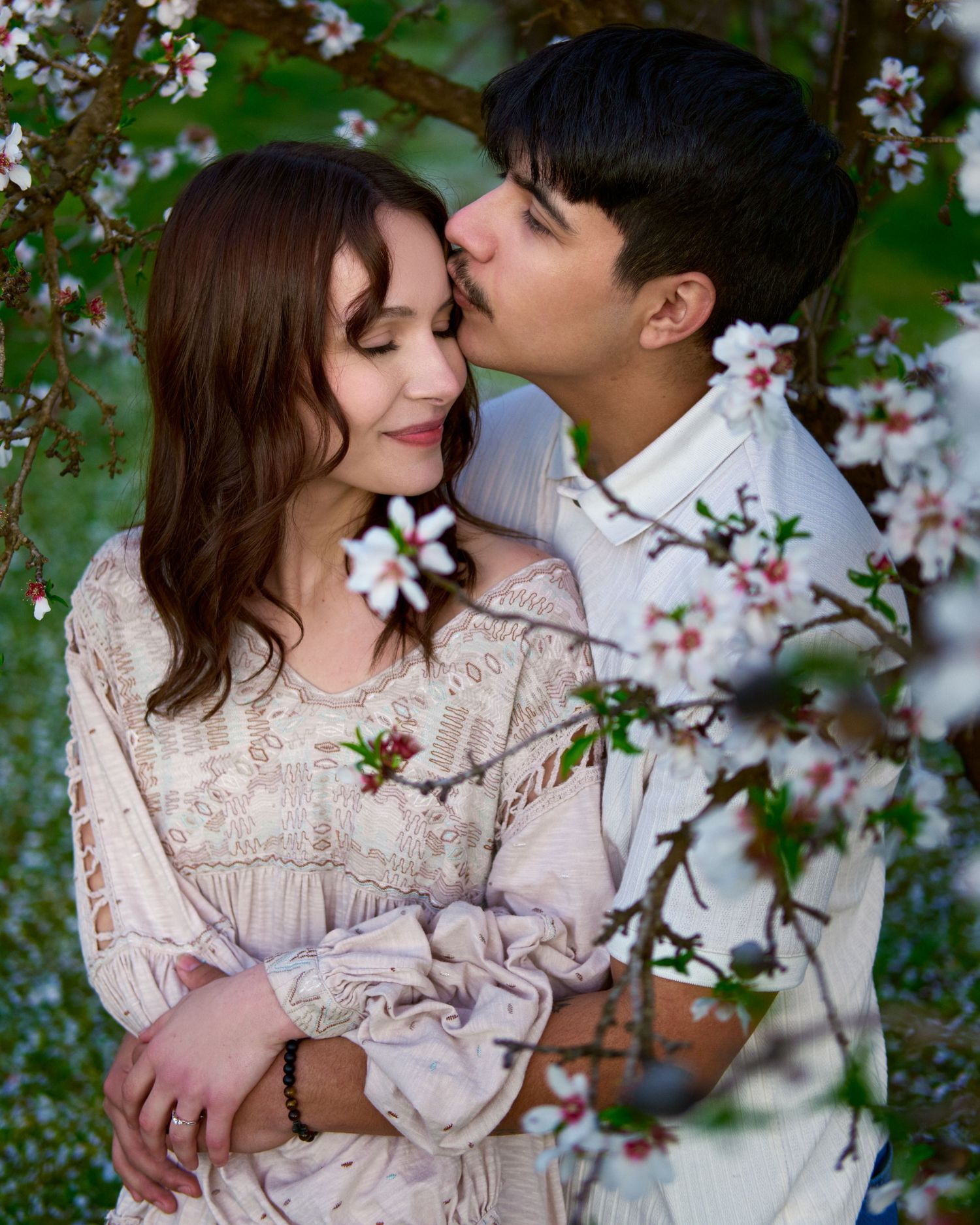 A couple embraces beneath blossoming spring branches as a man gently kisses the forehead of a woman in a cream lace blouse.