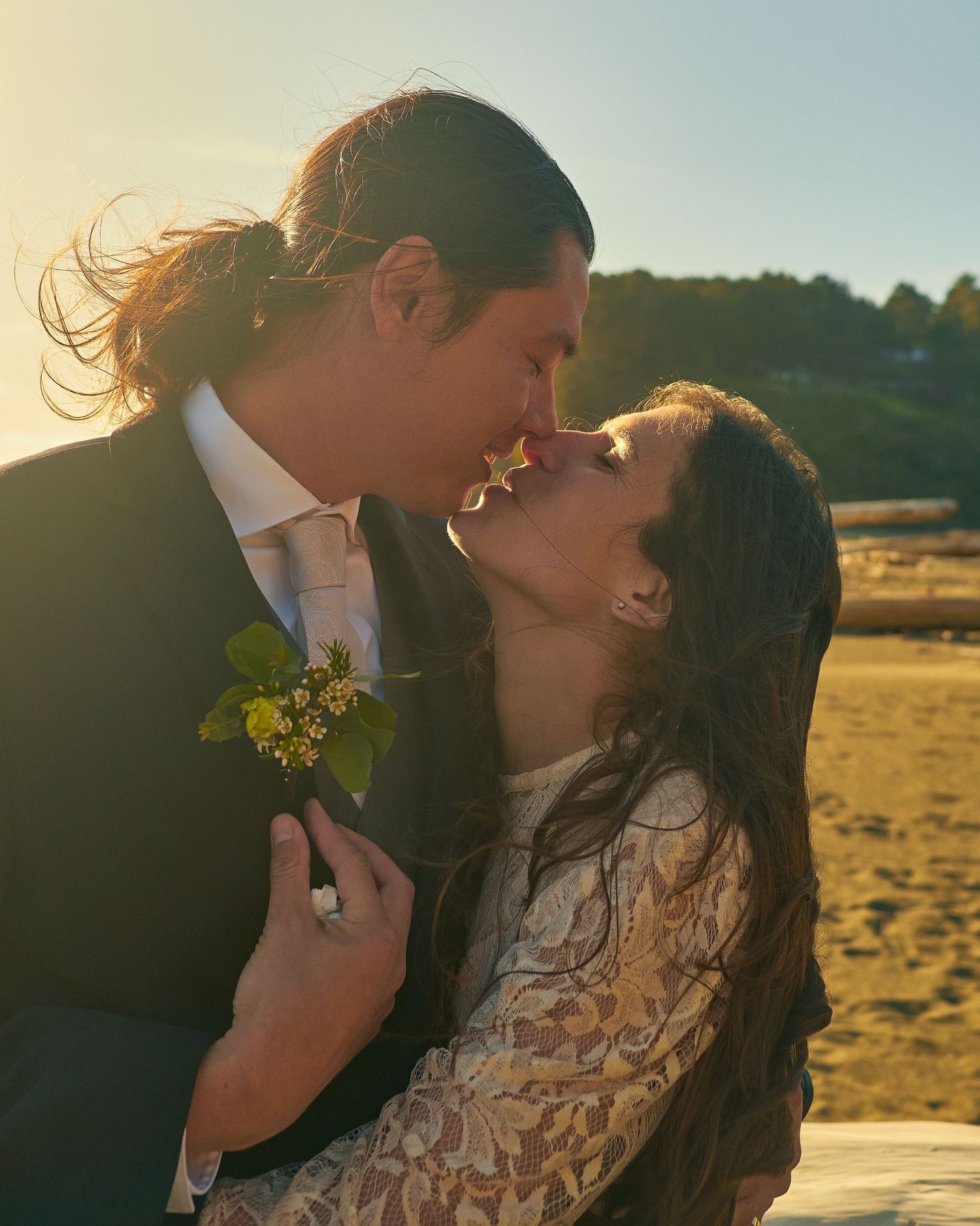 Couple kisses on a sunlit beach at sunset, the groom wearing a yellow floral boutonniere and the bride in a lace dress.