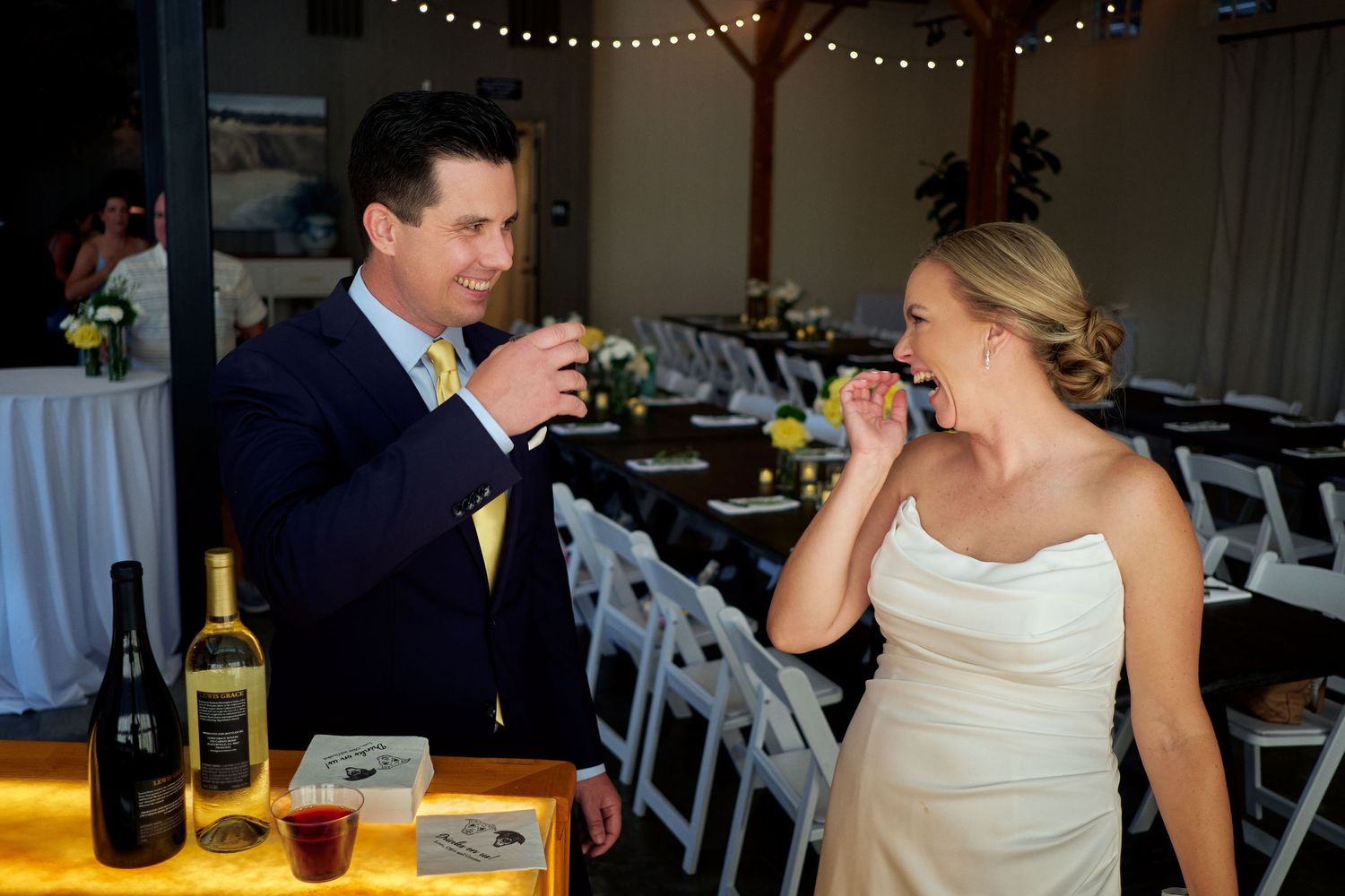 Smiling groom in a navy suit and bride in a white gown laugh while sampling wine at a festive reception with string lights and tables.