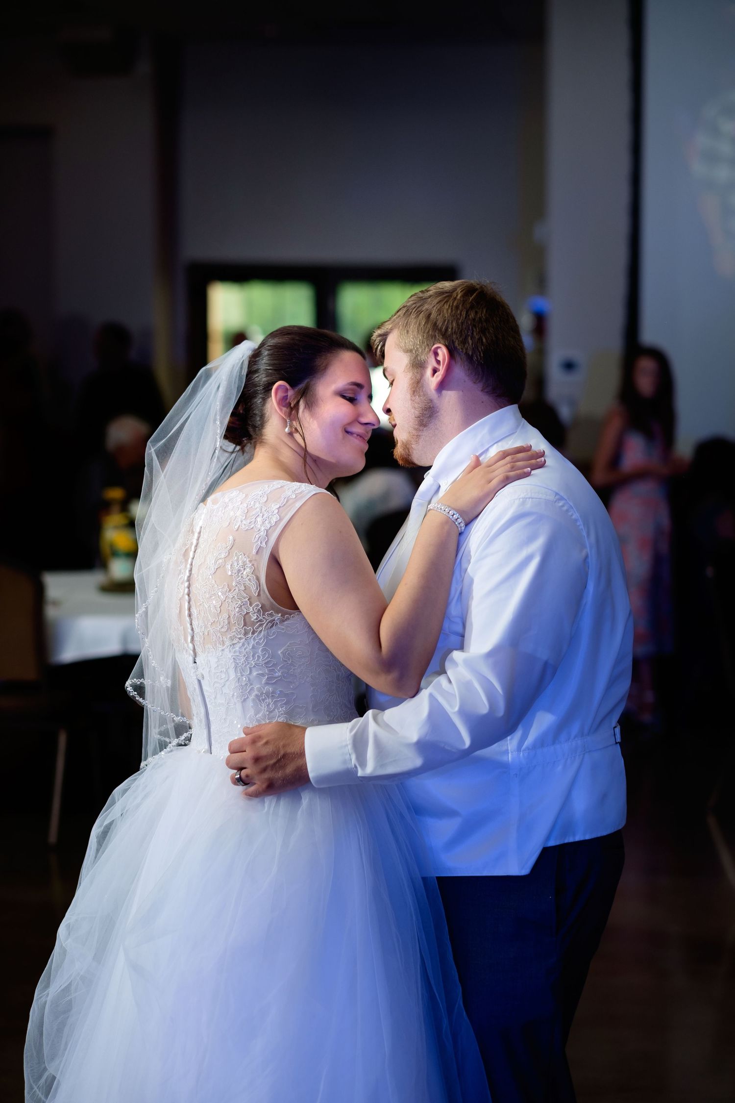 Bride in a lace gown and veil shares a tender first dance with her groom in a white shirt at an indoor wedding reception.