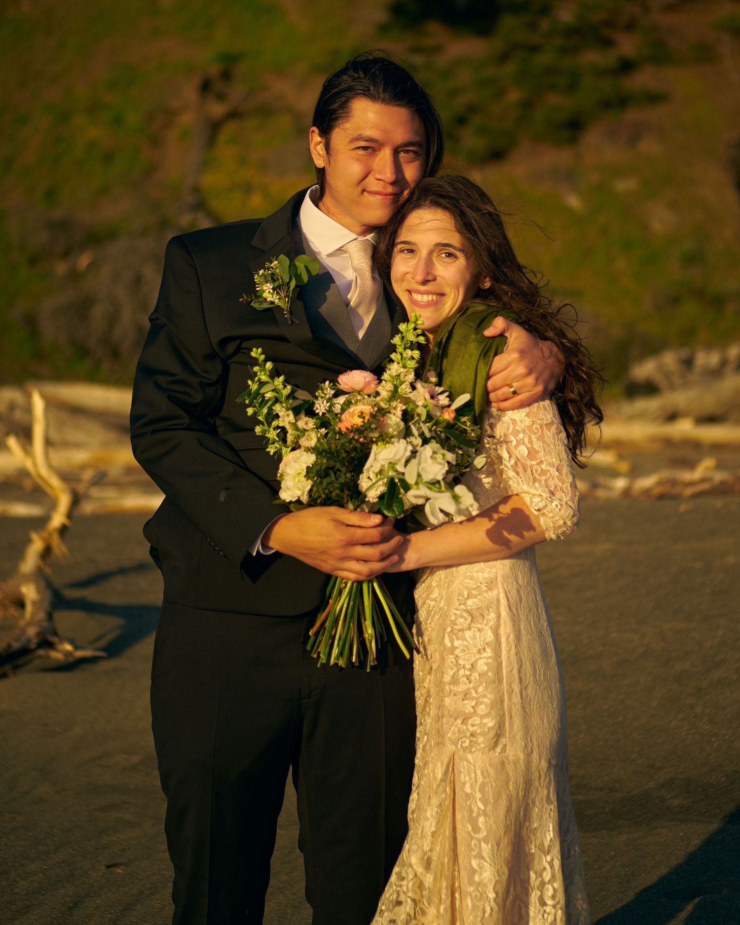 Smiling bride in a lace gown and groom in a dark suit embrace on a beach at golden hour, holding a white and pink floral bouquet.