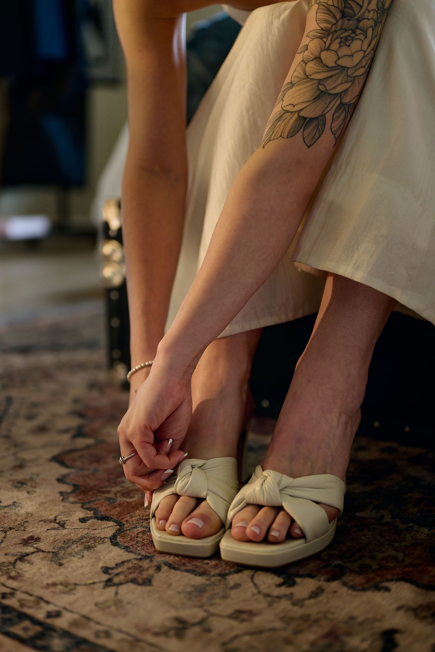 A woman with a floral arm tattoo adjusts her cream knotted platform sandal while wearing a white dress on a patterned rug.