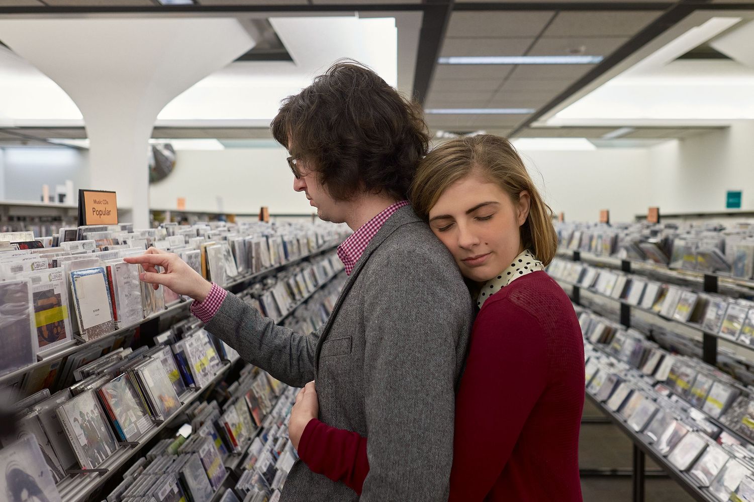 A woman in a red sweater hugs a man from behind as he browses CDs in a music store with shelves of albums.