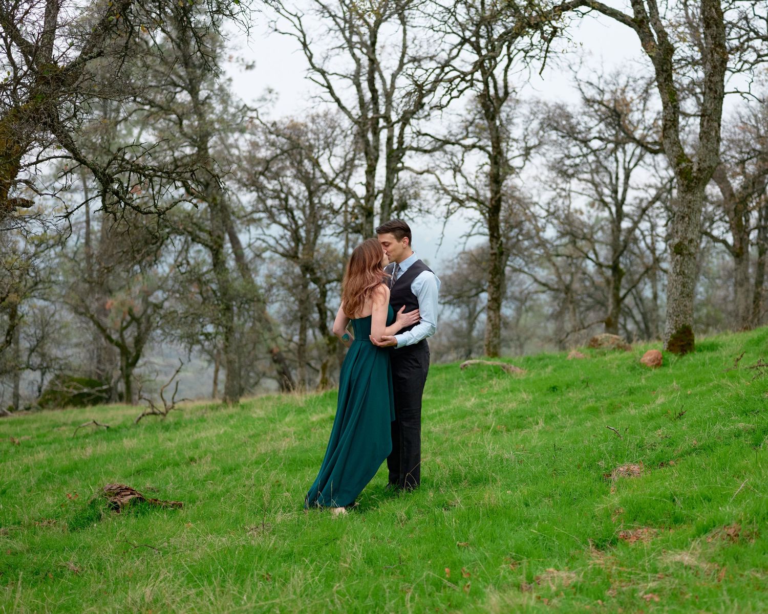 A couple in a dark suit and long green dress embrace on a grassy hillside surrounded by bare oak trees on a misty day.