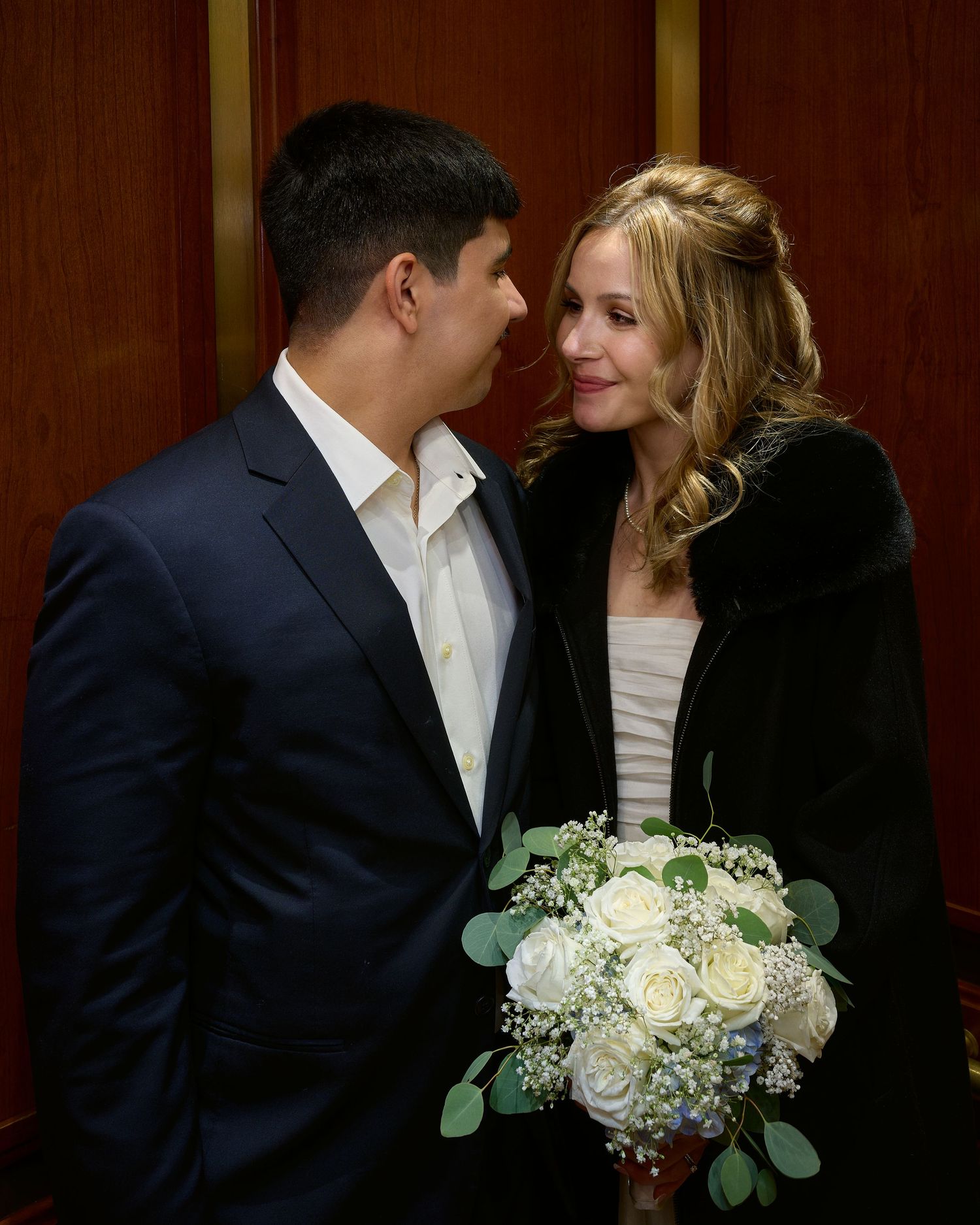 A smiling woman in a black fur wrap holds a white rose bouquet while gazing at a man in a navy suit in a warm wood-paneled room.