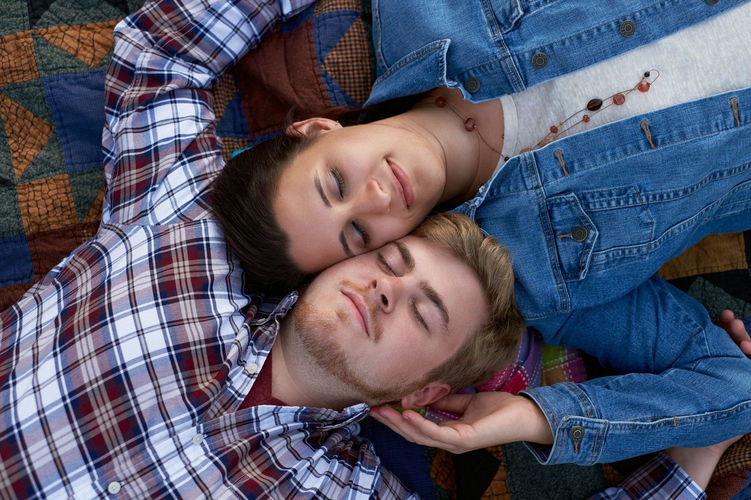Two young men lie close together with eyes closed, one in a plaid shirt and one in a denim jacket, resting on a colorful quilt.