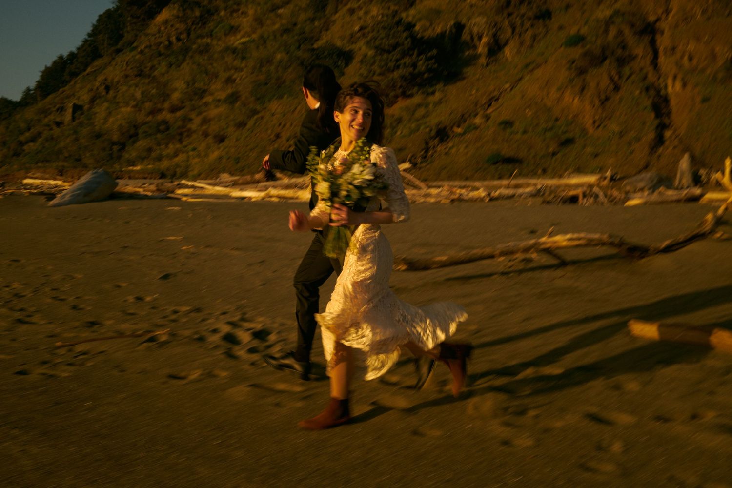 A bride in a white dress holds a bouquet while running along a sunlit beach with her partner beside rocky cliffs.