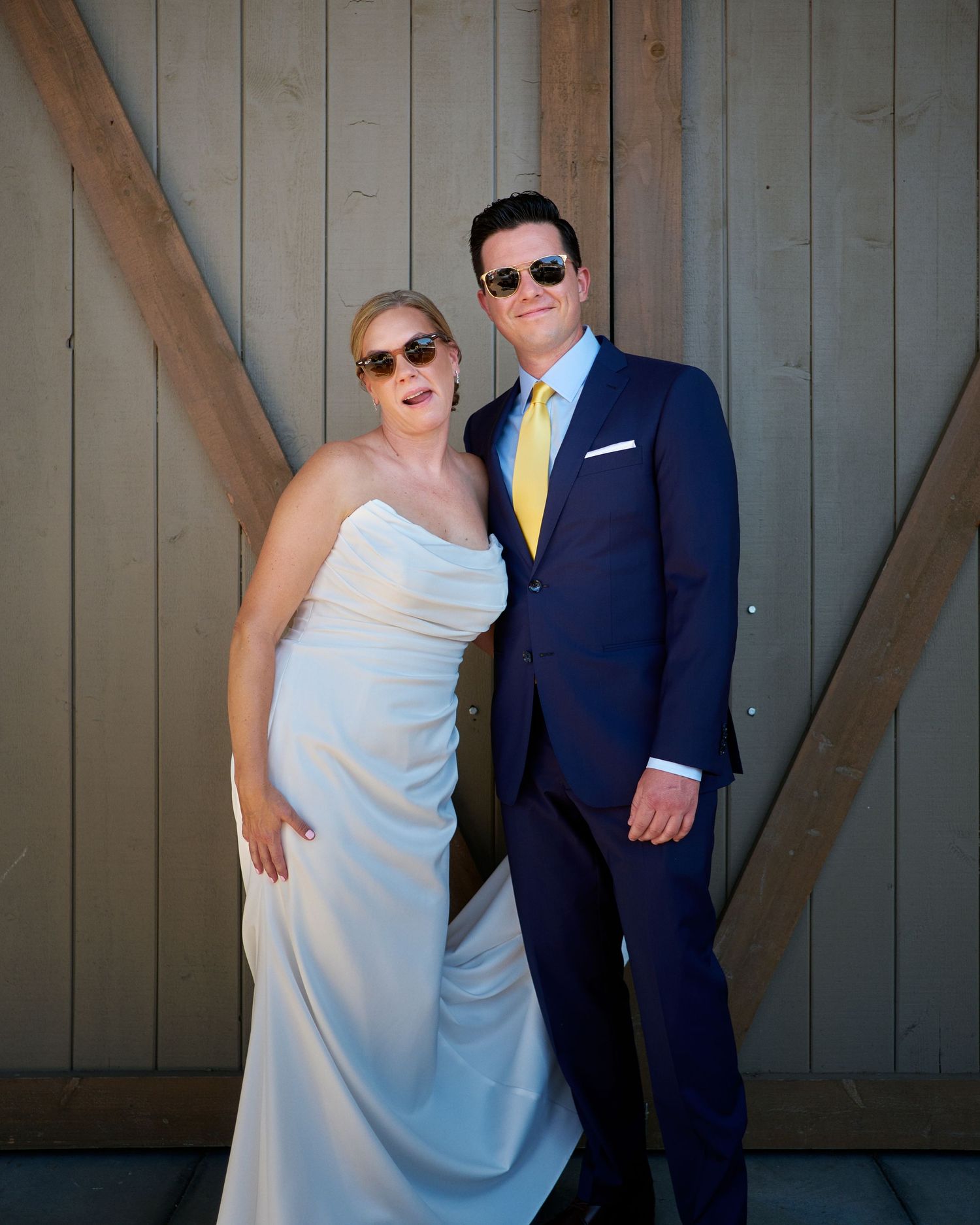 Smiling bride in a white strapless gown and groom in a navy suit and yellow tie pose in sunglasses before a rustic barn door.