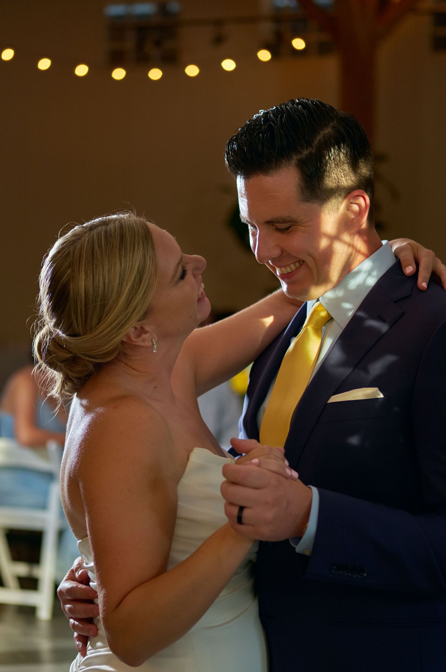 Smiling bride and groom in a navy suit with a yellow tie slow dance under string lights at their wedding reception.