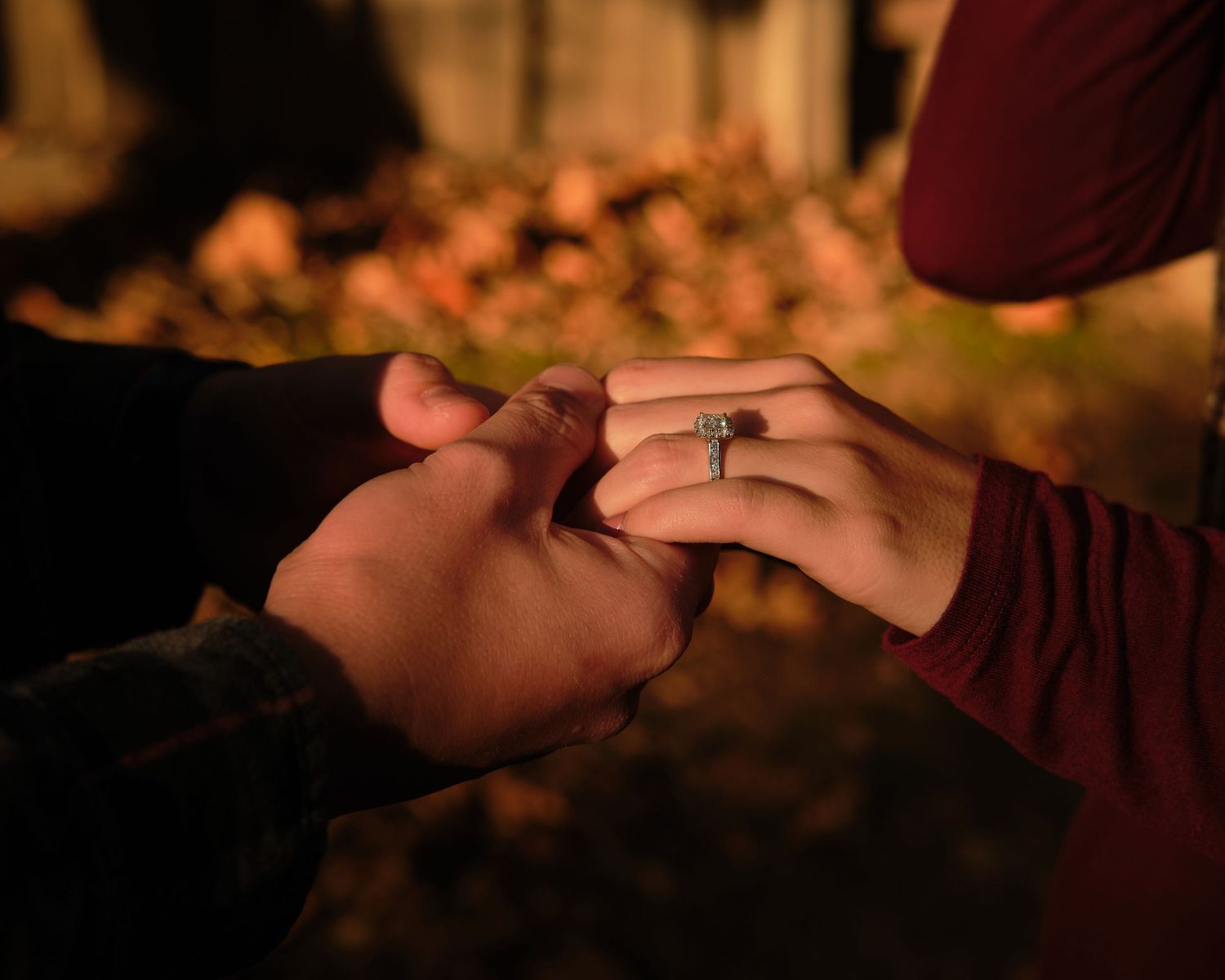 Close-up of a person holding another's hand to show a sparkling engagement ring, with warm autumn leaves softly blurred behind.