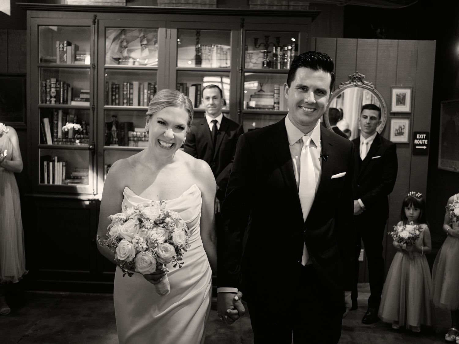 A smiling bride holding a pink rose bouquet walks hand in hand with her groom in a black and white wedding photo.