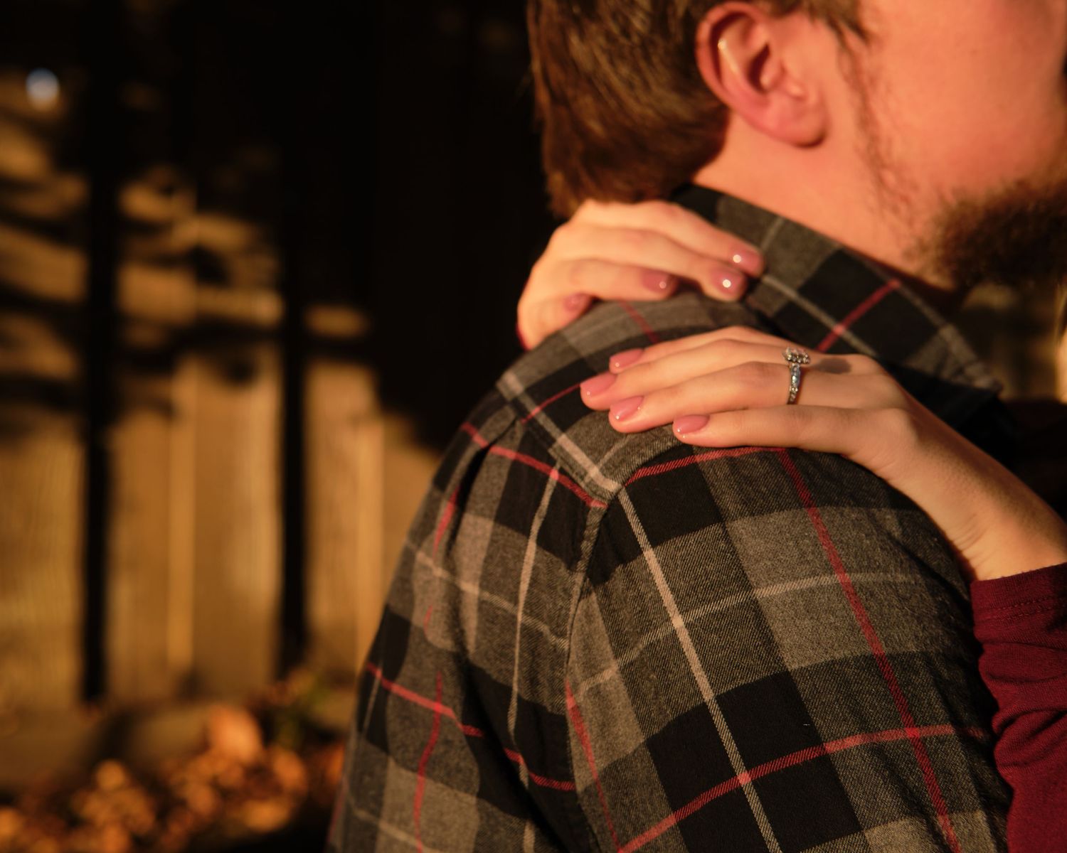 A woman's hand with a ring rests on the shoulder of a man in a gray and red plaid flannel shirt in warm evening light.