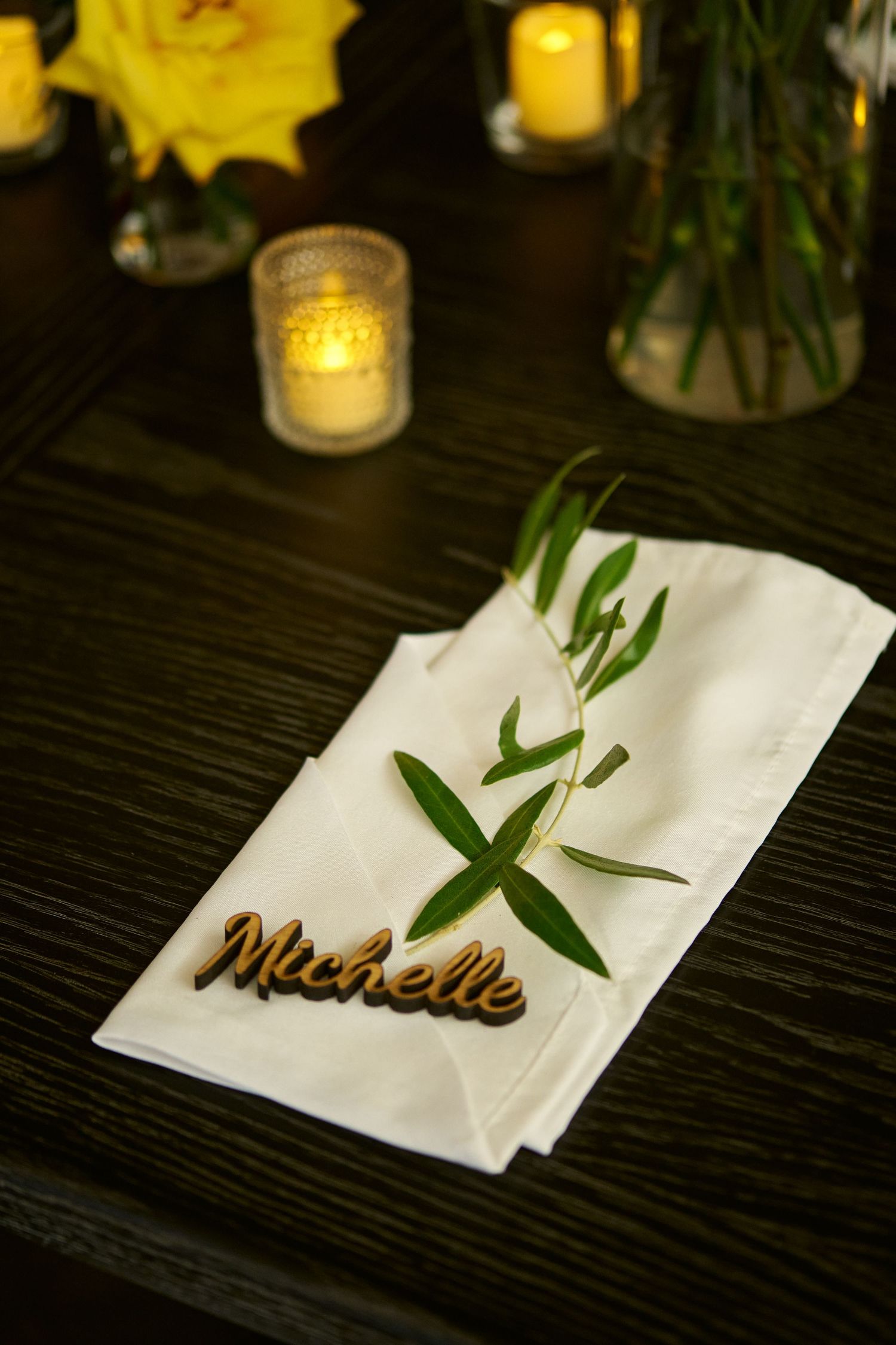 A wooden laser-cut Michelle place card rests on a white linen napkin with an olive sprig, beside candles and yellow roses.