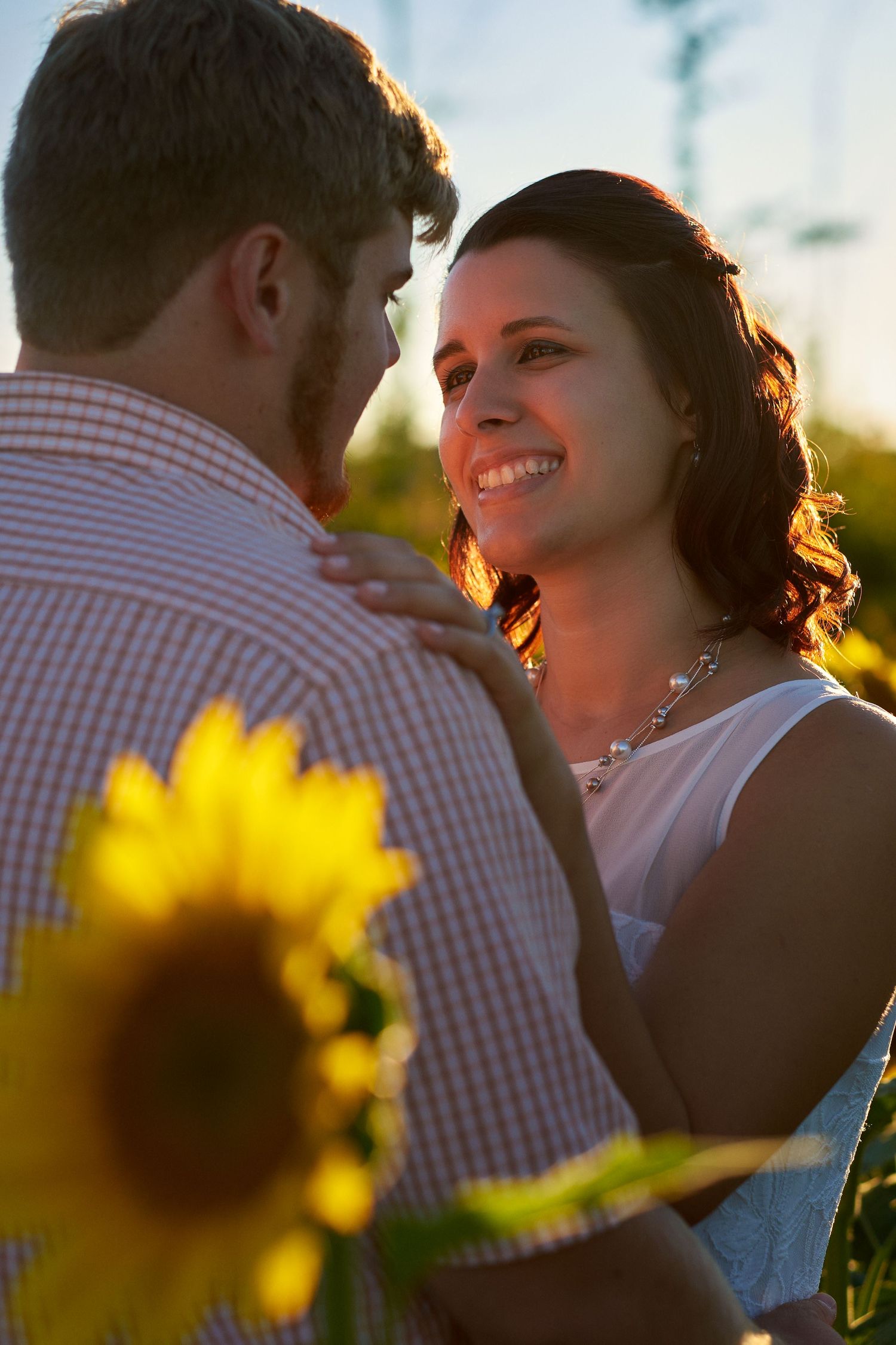 A smiling woman in a white top and necklace looks at a man in a checkered shirt in golden sunset light near a sunflower.